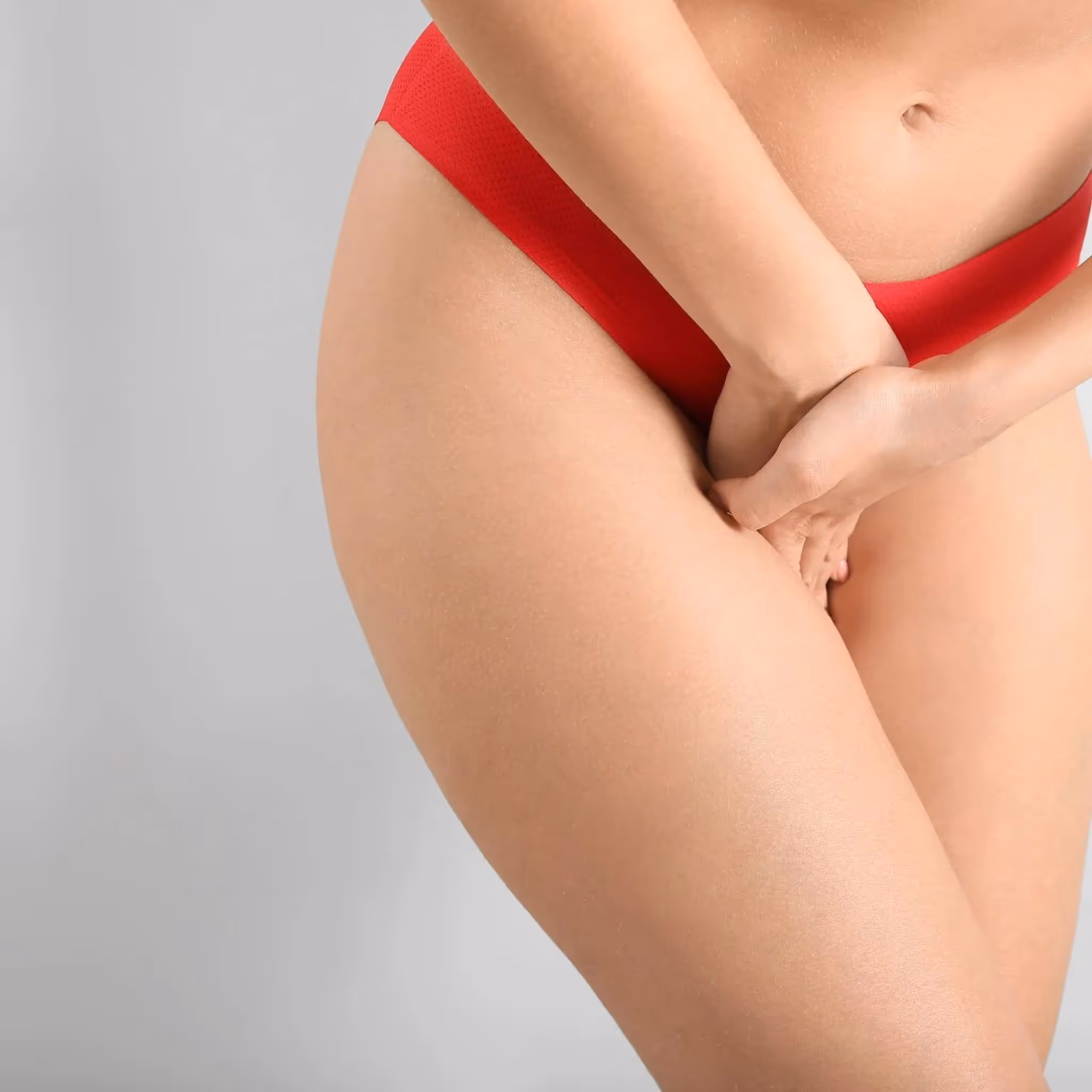 En person som har på seg rødt undertøy står med bena litt fra hverandre, og hendene dekker bekkenområdet mot en vanlig grå bakgrunn.