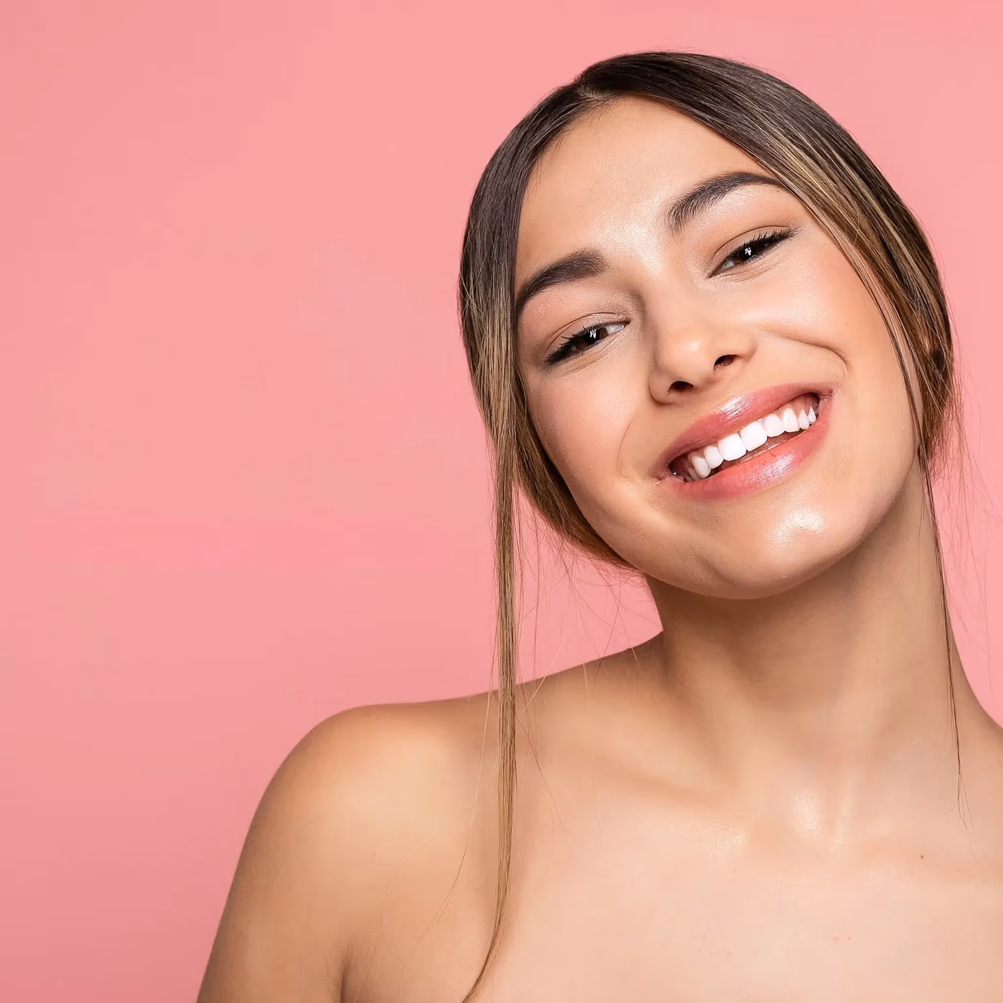En kvinne med rett brunt hår på midten smiler bredt mot en rosa bakgrunn. Hun har på seg minimalt med sminke og skuldrene er nakne.