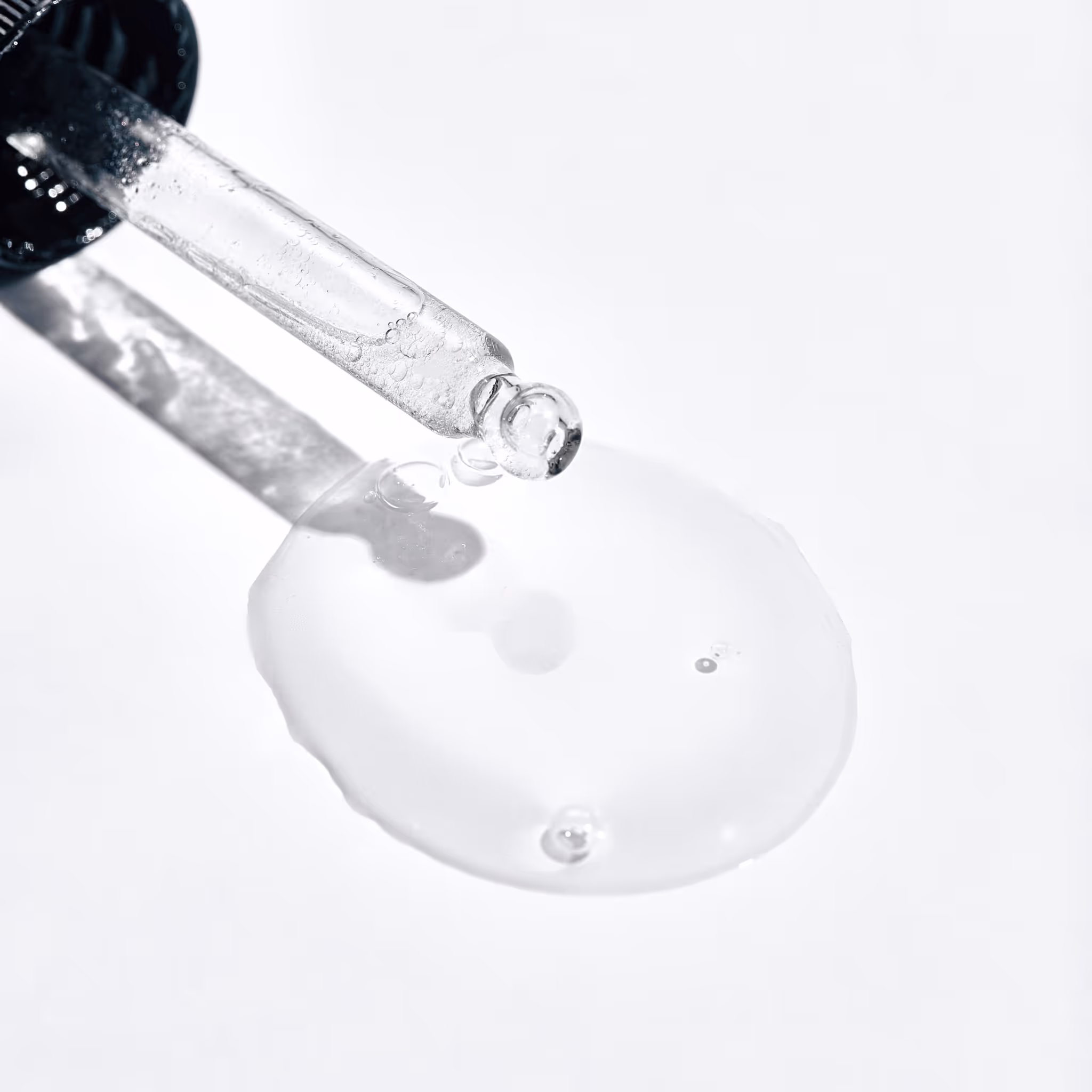 Nærbilde av en klar væske som dispenseres fra en glassdråpe på en hvit overflate. Dropperen vippes for å frigjøre væsken, som danner et lite basseng med synlige dråper rundt seg.