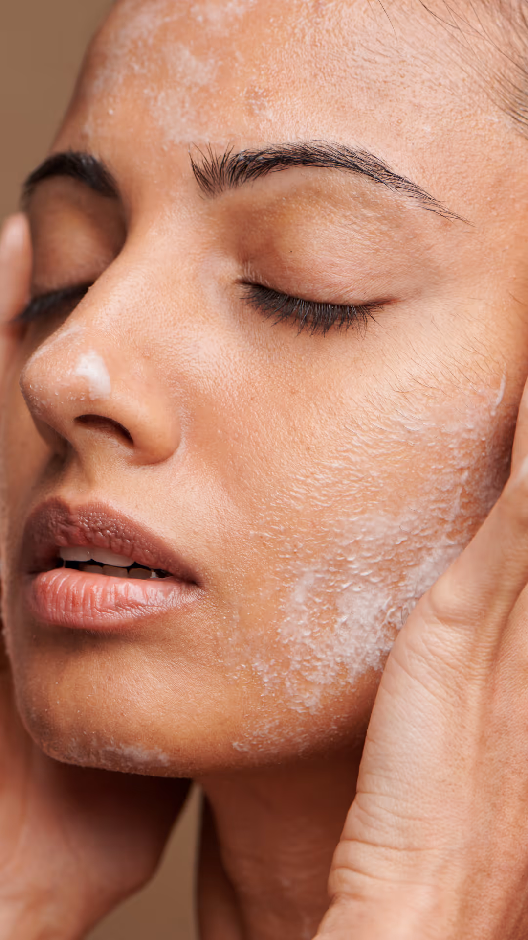 Nærbilde av en person som forsiktig vasker ansiktet med lukkede øyne, med en skummende rengjøringsmiddel på huden. Hendene deres masserer rensemidlet inn i kinnene, og gir en følelse av avslapning og hudpleierutine.