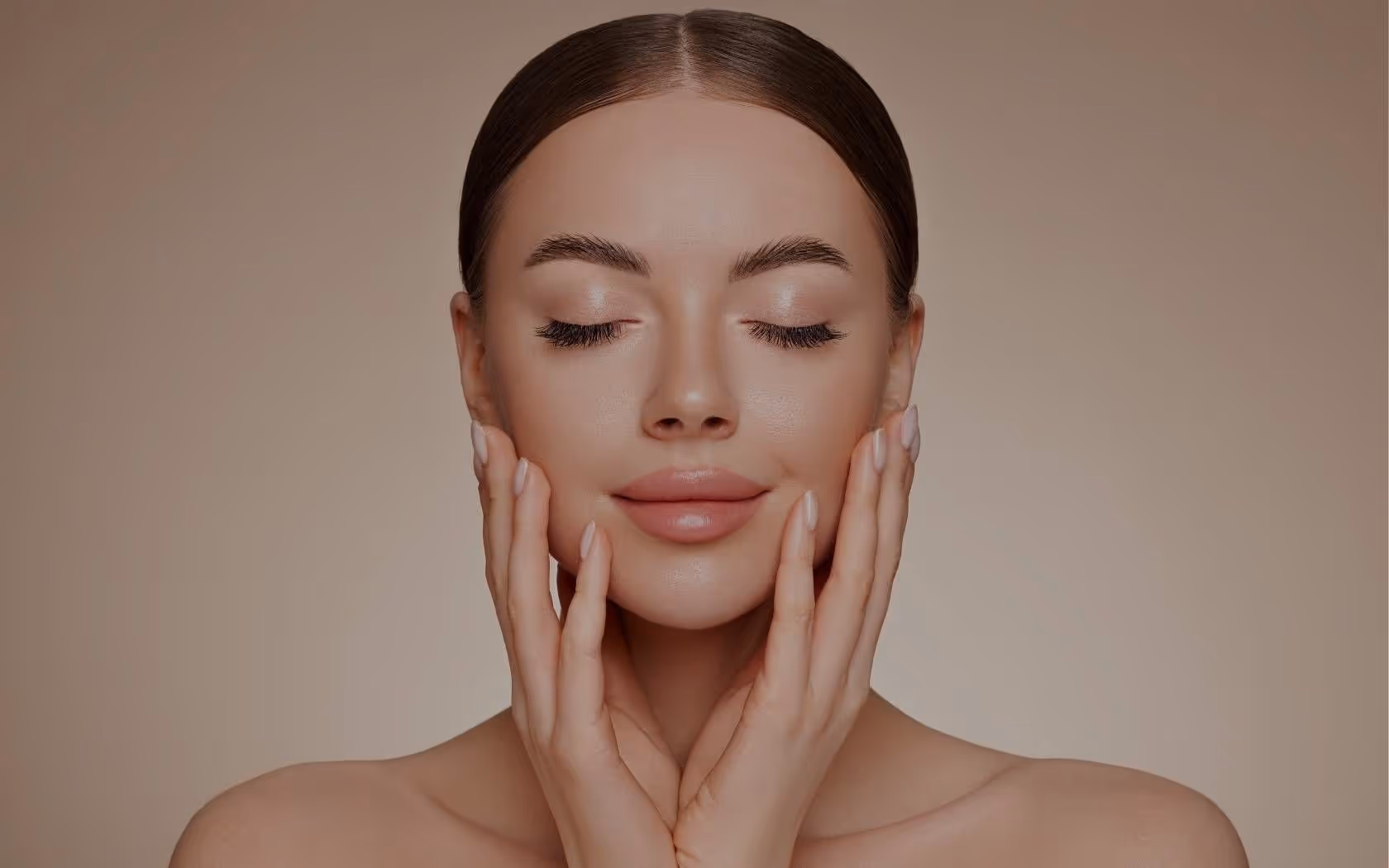 En kvinne med et rolig uttrykk, lukkede øyne, berører ansiktet hennes forsiktig med hendene mot en nøytral bakgrunn.