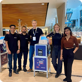 Five smiling people standing indoors around a blue and white kiosk with a digital screen, inside a modern building with wooden ceiling and glass walls.