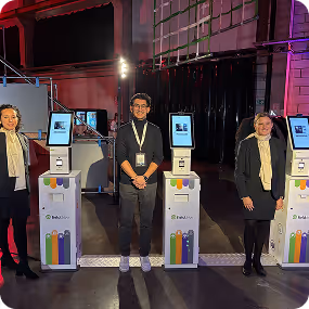 Three people standing indoors beside colorful self-service kiosks with screens, in a dimly lit room.