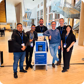 Group of six people standing around a blue support cart inside a modern building.