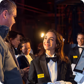 Female event staff in a tuxedo smiling and interacting with guests at a registration or ticketing desk.
