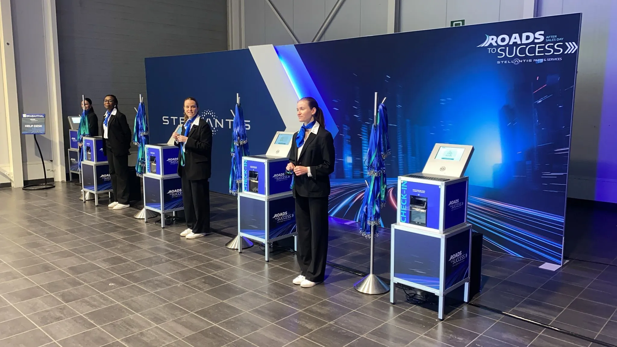 Four women in black suits and blue scarves standing behind check-in kiosks at a Roads to Success event with a blue-themed background.