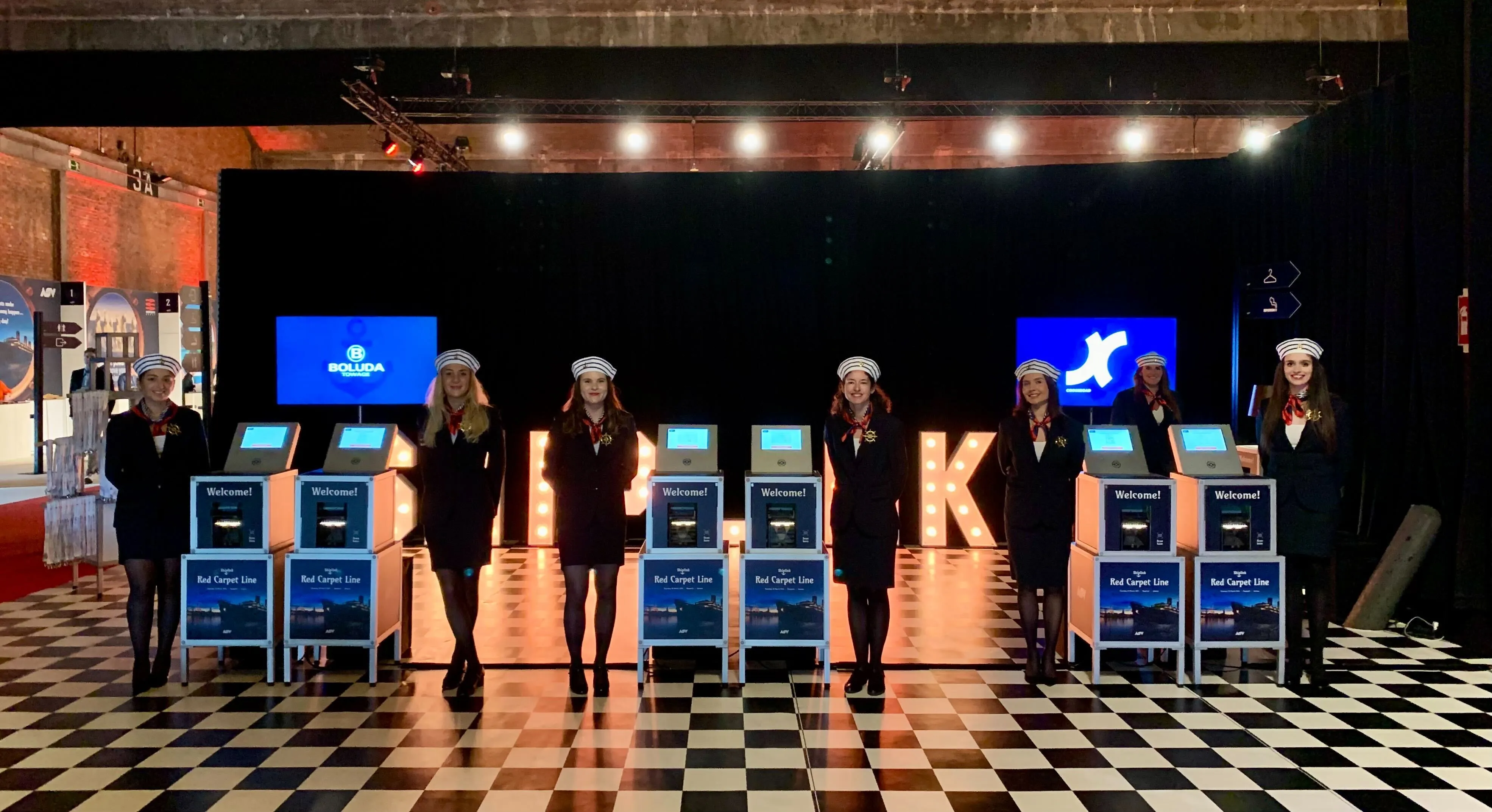 Seven women in navy uniforms and striped hats standing behind check-in kiosks on a black and white checkered floor with illuminated letters spelling 'BILBAO'.