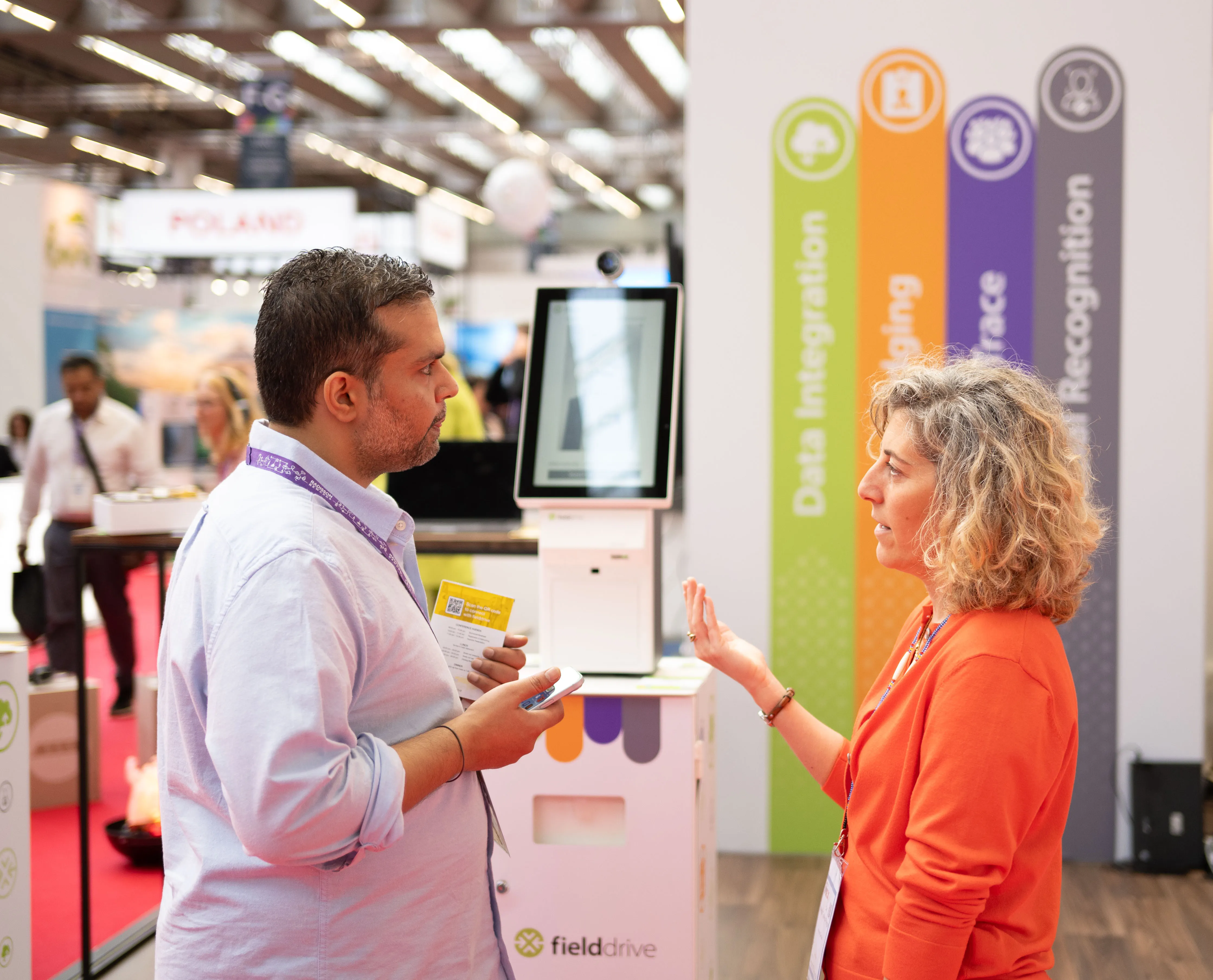 Two people engaged in conversation at a trade show booth with a fielddrive kiosk in the background.