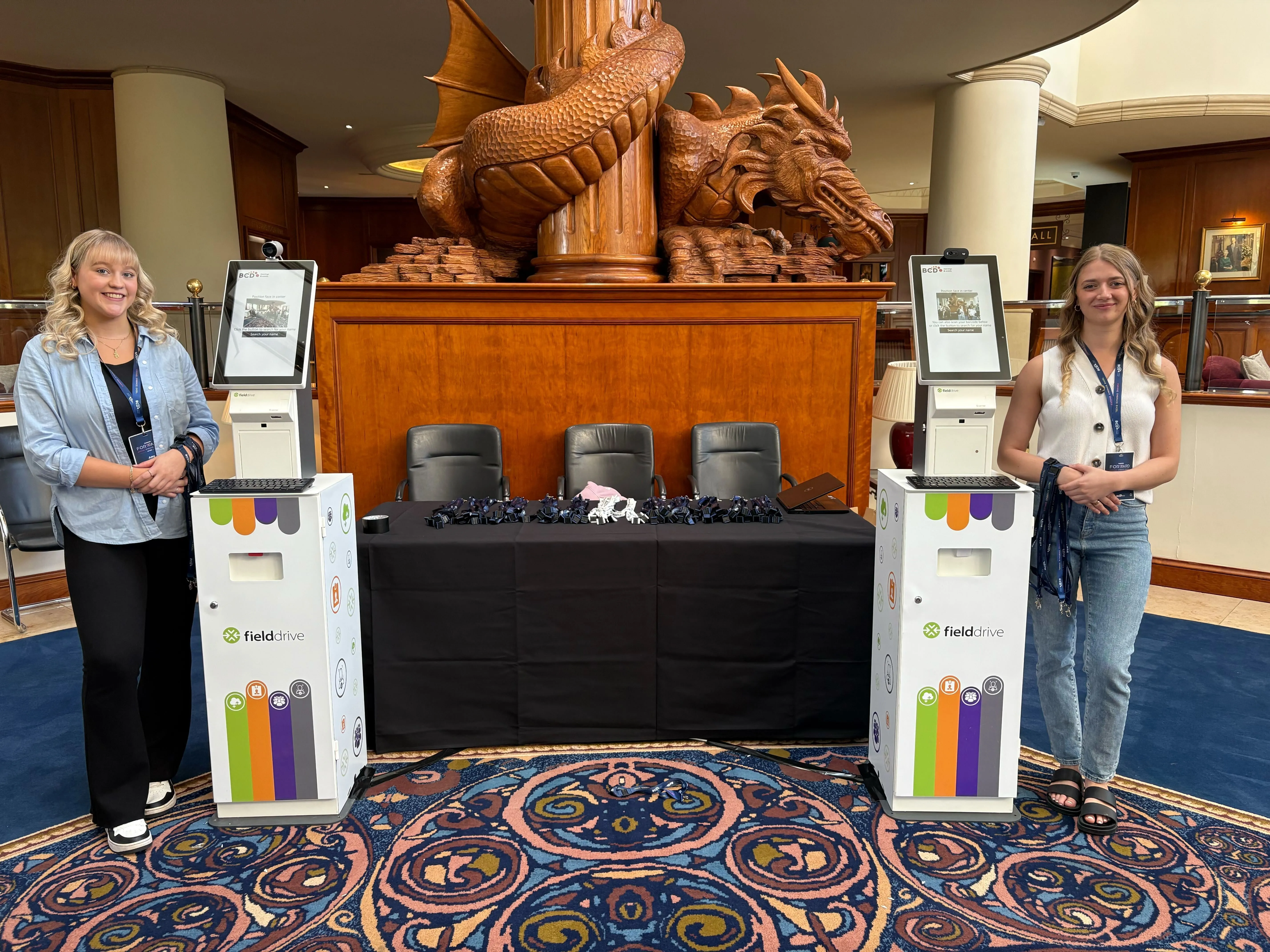 Two women standing on either side of a check-in table with Fielddrive kiosks and a carved wooden dragon sculpture in the background.