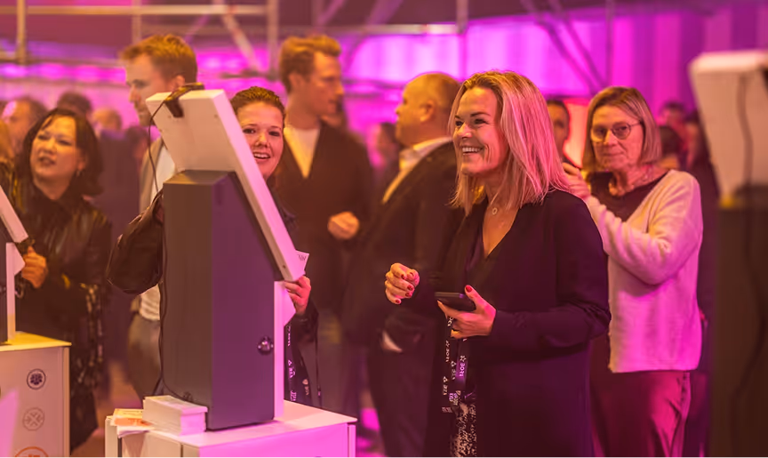 People interacting and smiling near digital kiosks at an event with purple lighting.