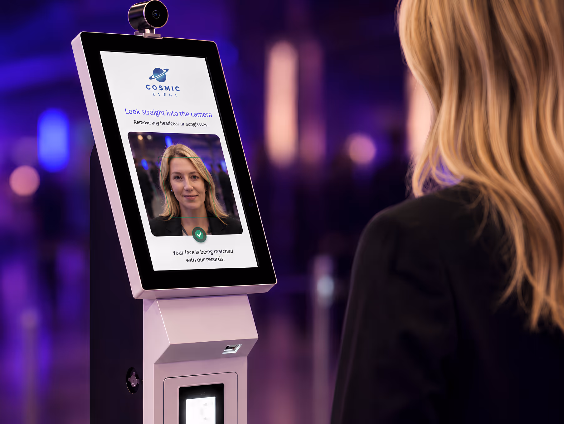 Woman using a facial recognition kiosk with instructions to look straight into the camera for event check-in.
