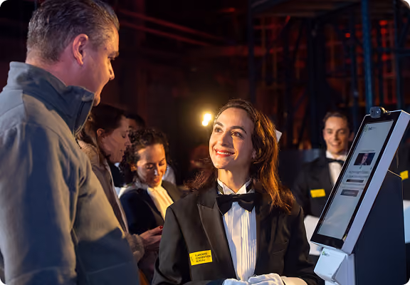 Woman in tuxedo greeting a man at a check-in kiosk with other formally dressed staff nearby.
