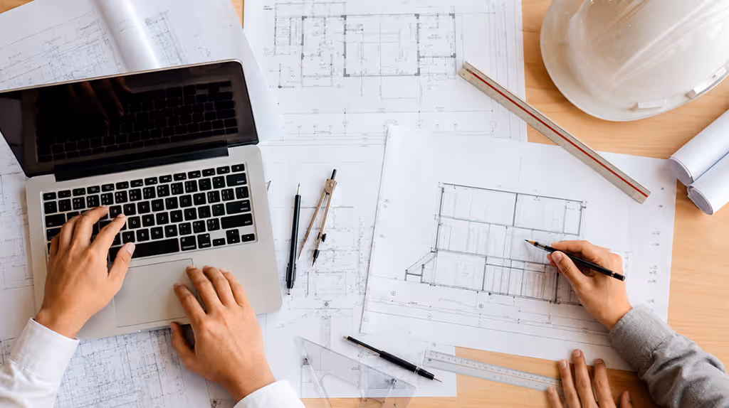 Dois profissionais trabalham juntos sobre plantas técnicas em mesa de escritório, com laptop, capacete e instrumentos de desenho, representando a atuação conjunta entre engenheiro e arquiteto na emissão de ART e RRT.