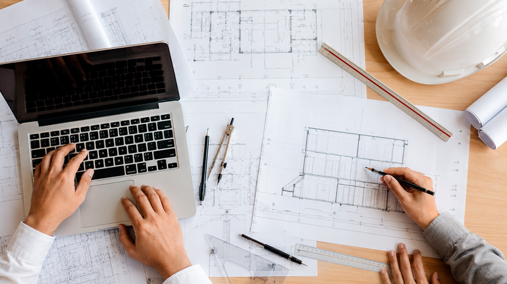 Dois profissionais trabalham juntos sobre plantas técnicas em mesa de escritório, com laptop, capacete e instrumentos de desenho, representando a atuação conjunta entre engenheiro e arquiteto na emissão de ART e RRT.