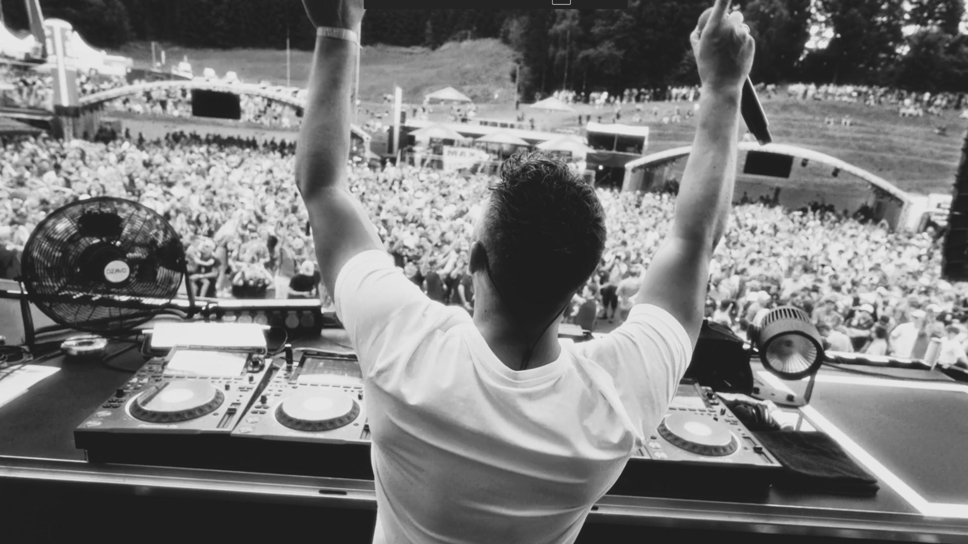 DJ with raised arms performing on stage in front of a large outdoor crowd.