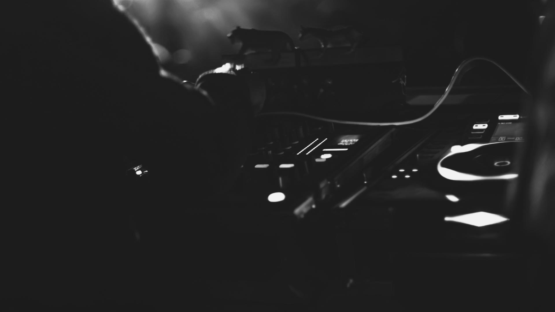Black and white image showing a DJ controller with illuminated buttons and knobs in low light.