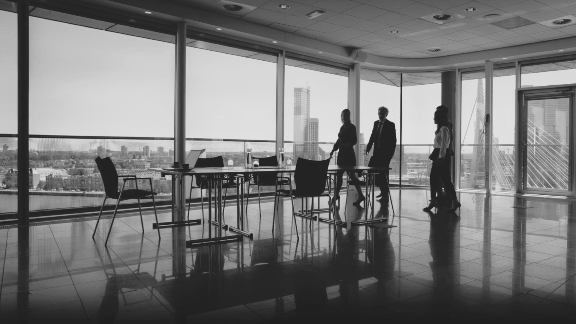 Silhouettes of four businesspeople walking past a conference table in a high-rise office with large windows overlooking a cityscape.