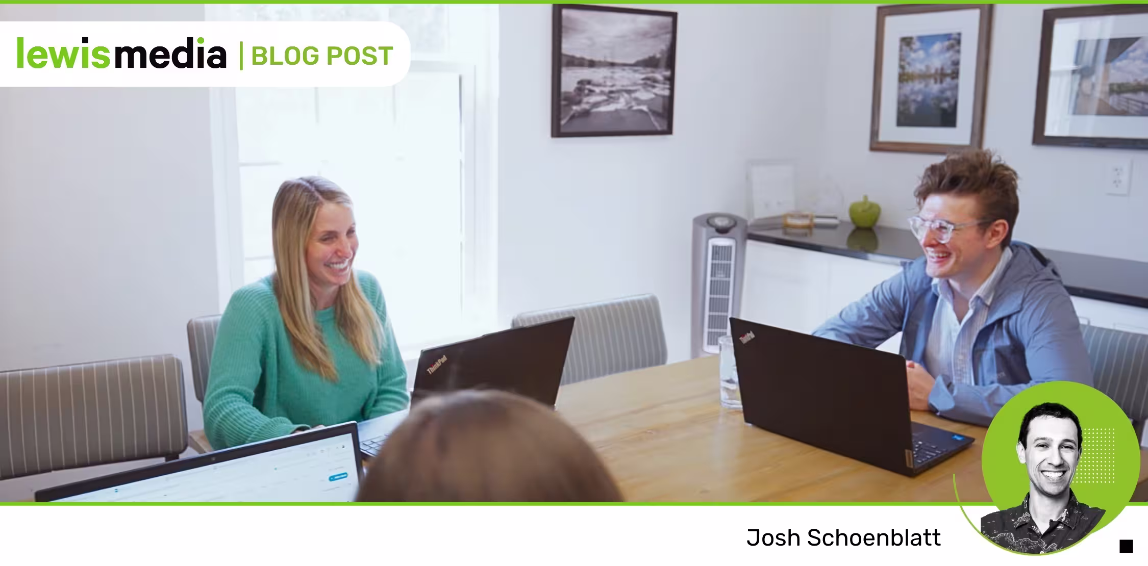 Two people smiling while sitting at a table with laptops in a bright office room.