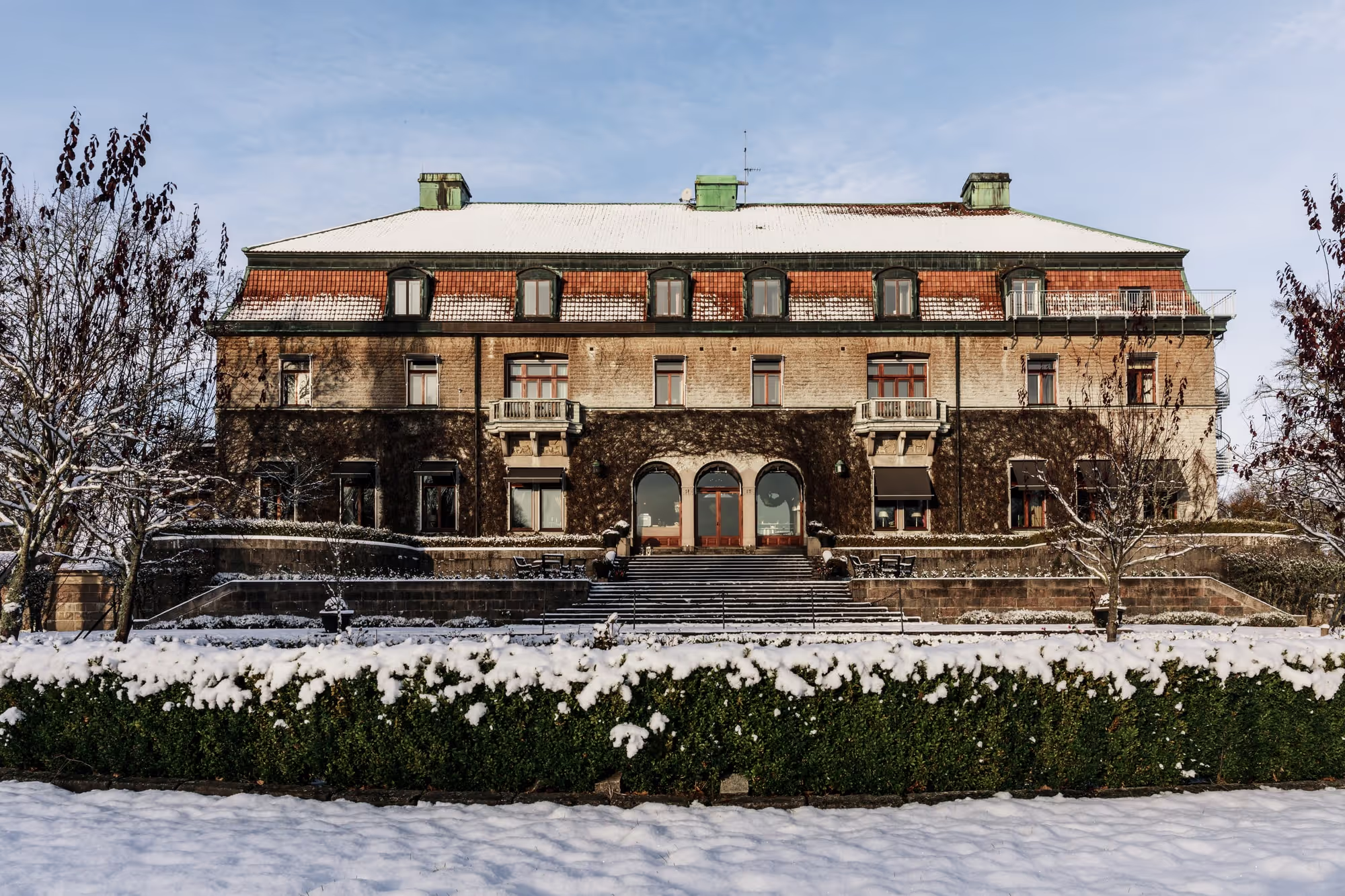 Stort historiskt tegelhus med snötäckt tak och trappa, omgiven av snö och kala träd.