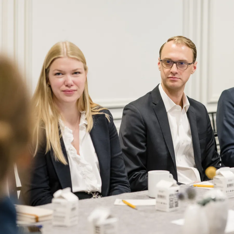 Two professionals, a woman and a man in business attire, seated at a meeting table with notebooks and cups.