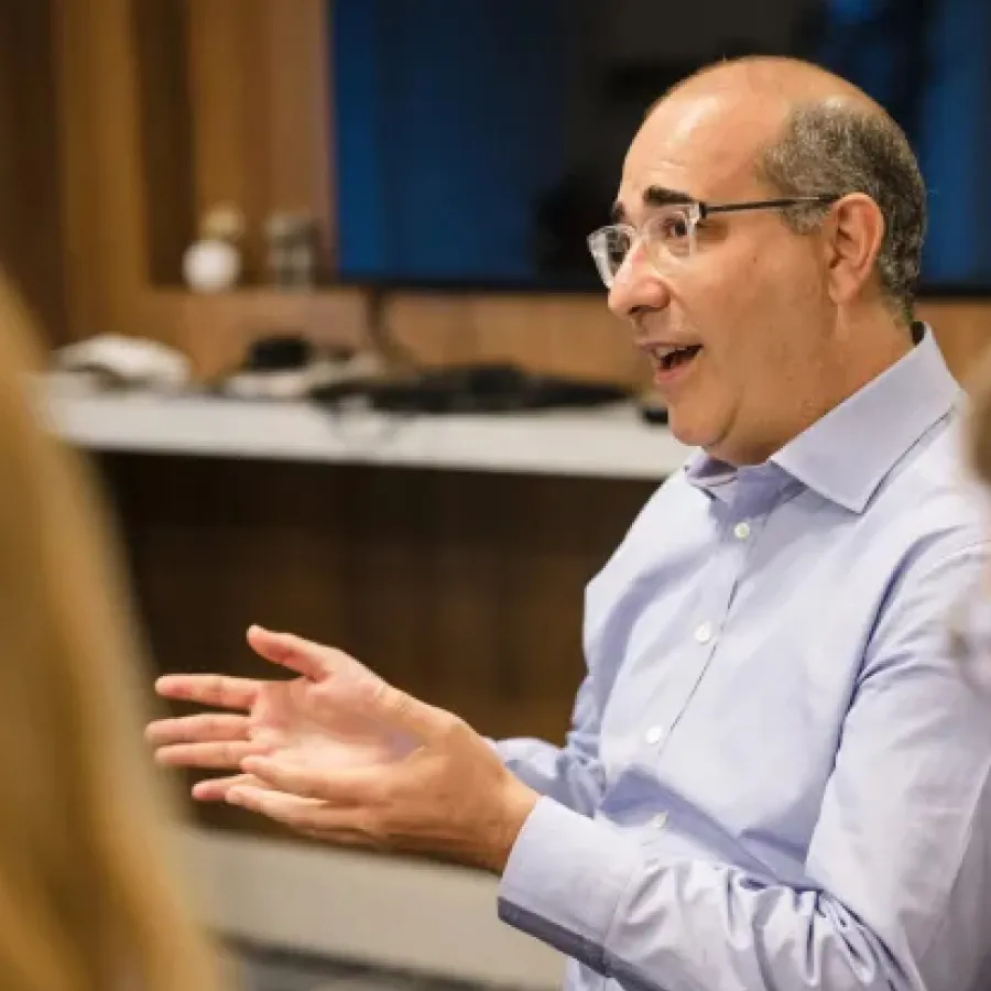 Man in glasses and light blue shirt speaking and gesturing with hands in an indoor setting.