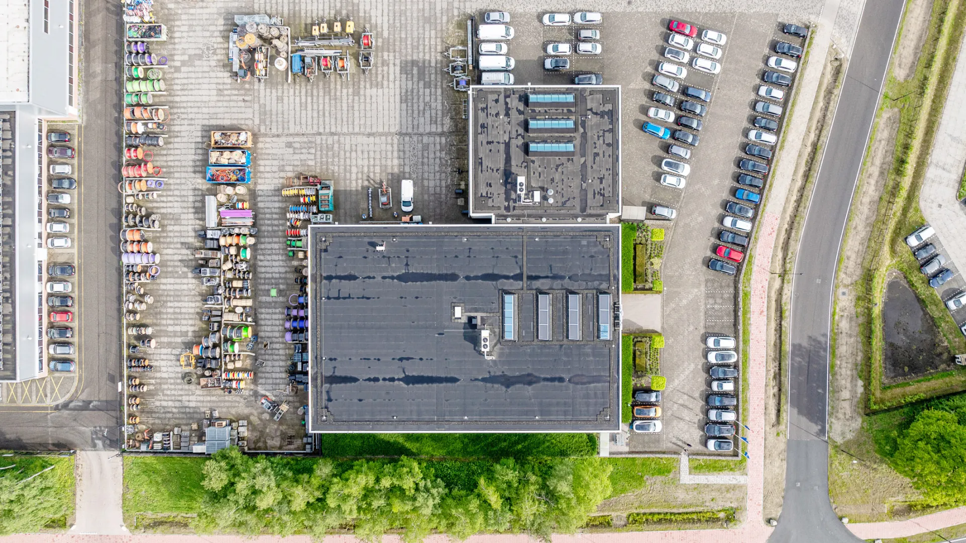Aerial view of industrial buildings with parked cars and organized outdoor storage of colorful barrels and materials.