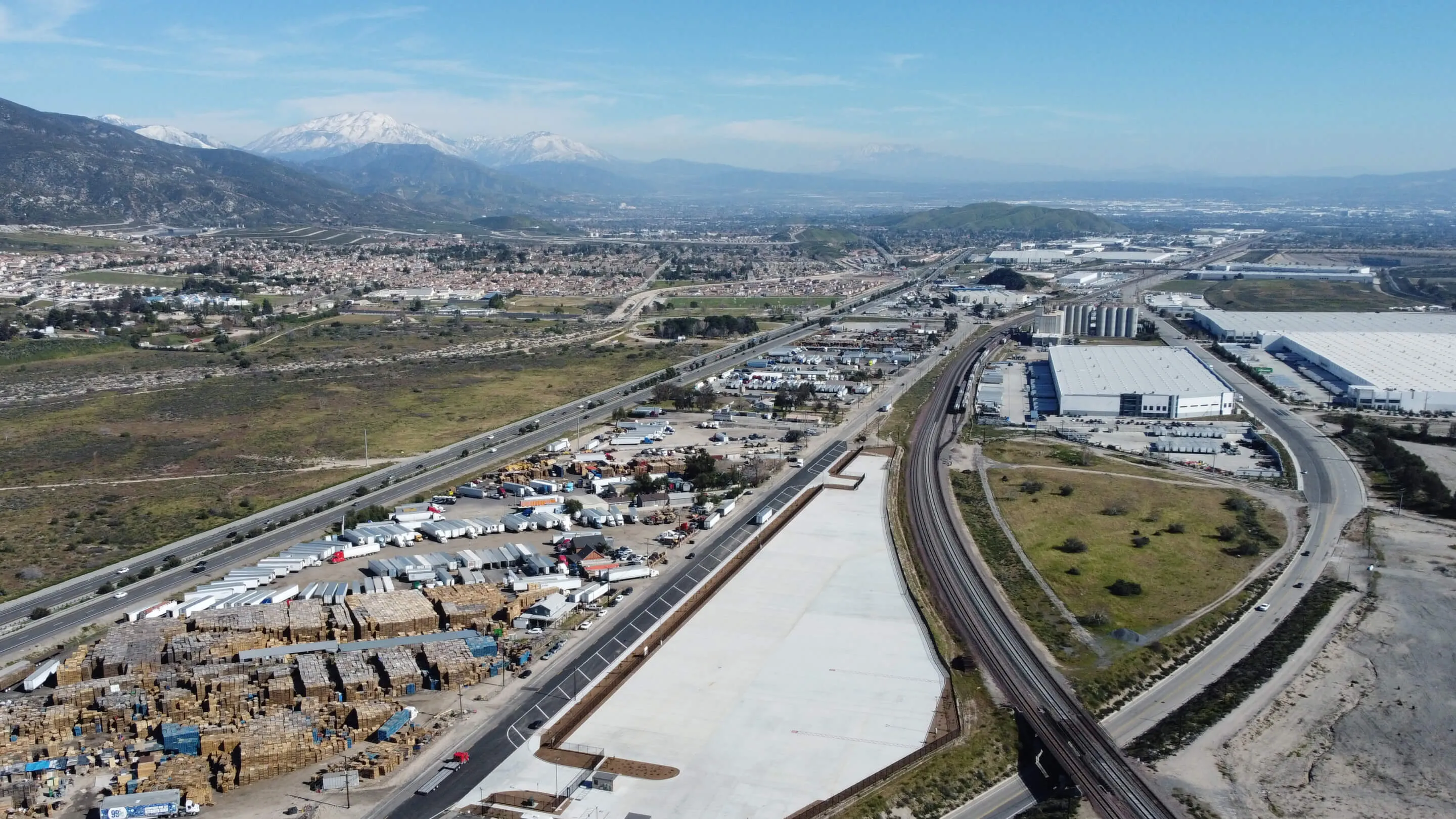 Aerial view of industrial area with warehouses, trucks, railroad tracks, and snow-capped mountains in background.