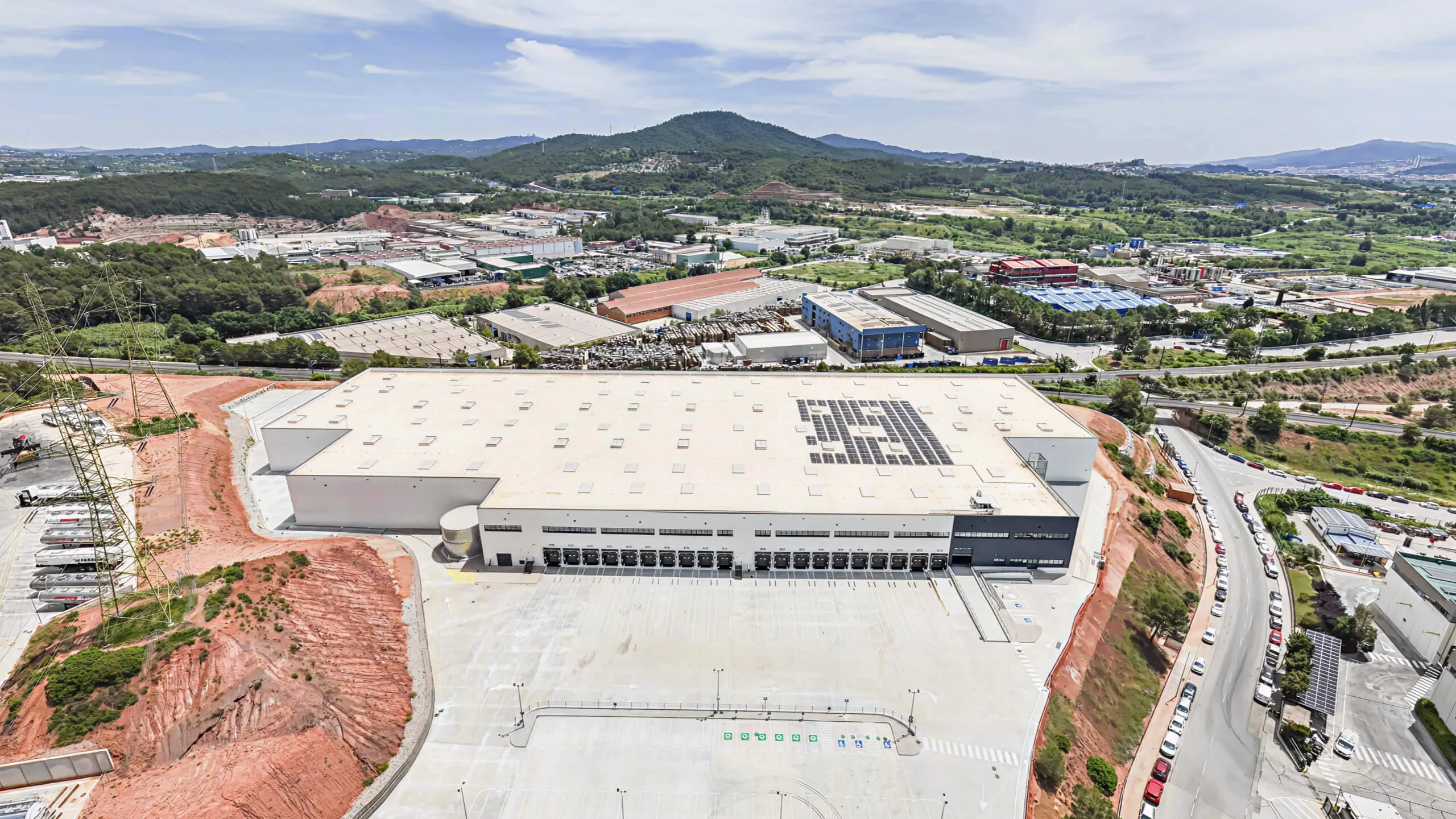 Aerial view of large warehouse with solar panels on roof and parking area surrounded by hilly landscape.