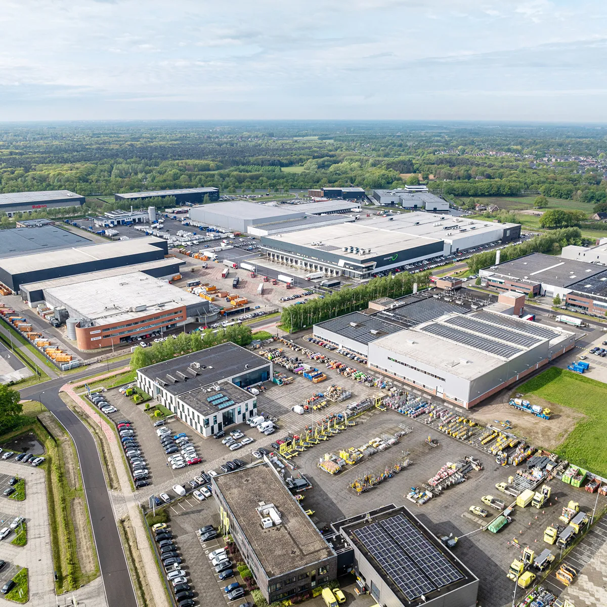 Aerial view of an industrial park with warehouse buildings, parked vehicles, and green landscape beyond.