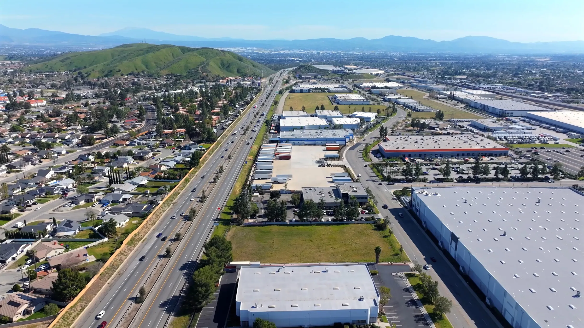 Aerial view of highway bordered by residential homes on left and industrial buildings on right beneath blue sky.