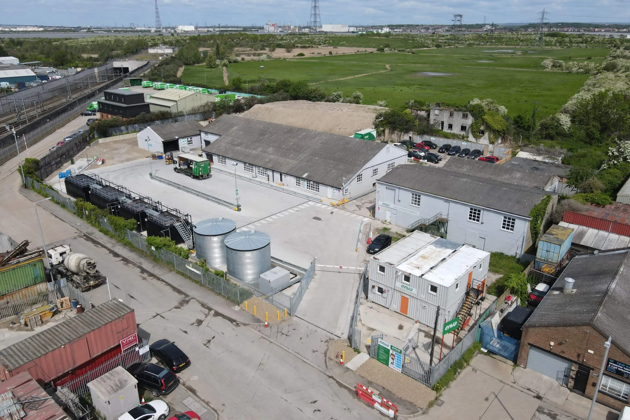 Aerial view of an industrial site with buildings, storage tanks, parked cars, and surrounding green fields.