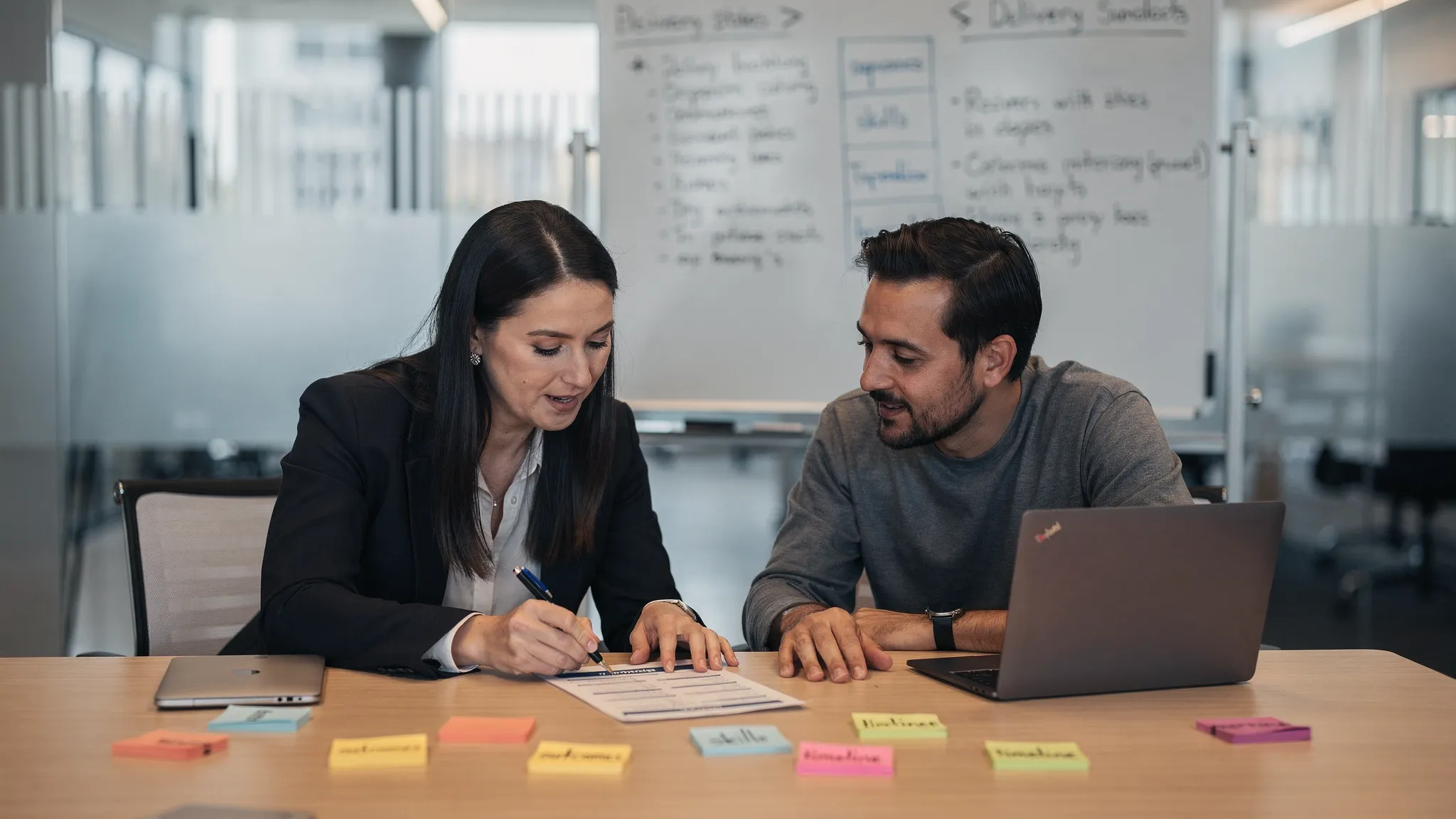 A hiring manager and a technical lead in a meeting room reviewing a role brief on paper, with sticky notes showing “outcomes”, “skills”, and “timeline”, and a whiteboard listing delivery milestones and interview stages.