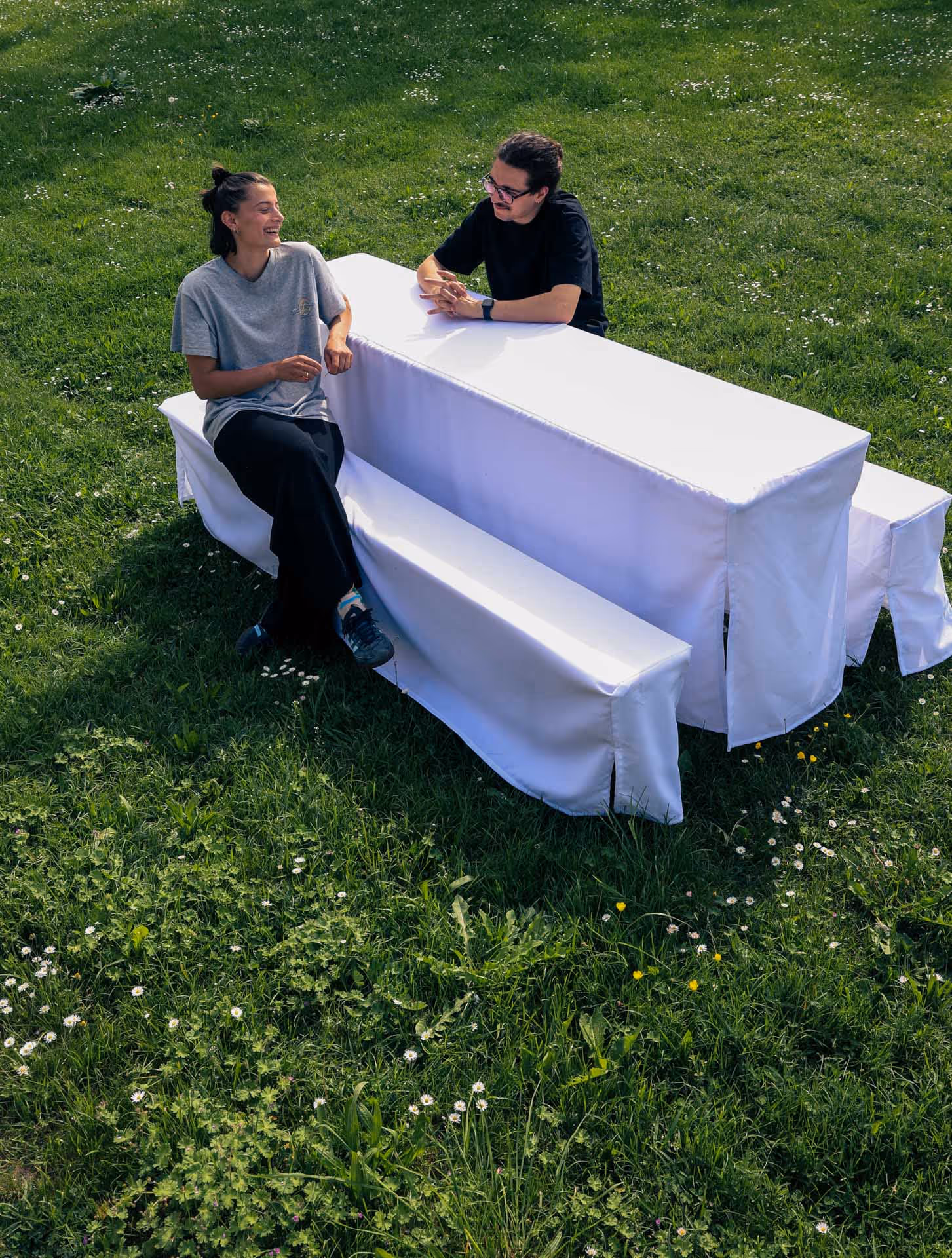 Bild zweier Personen, die auf einer Bierbankgarnitur gegenüber mit weißer, übergezogener Bierbankhusse sitzen. Die Bierbankgarnitur steht auf einer grünen Wiese.