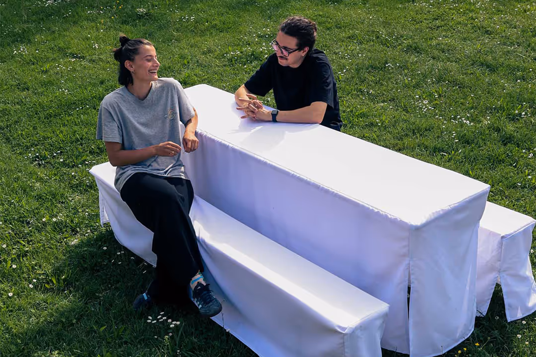 Bild zweier Personen, die auf einer Bierbankgarnitur gegenüber mit weißer, übergezogener Bierbankhusse sitzen. Die Bierbankgarnitur steht auf einer grünen Wiese.