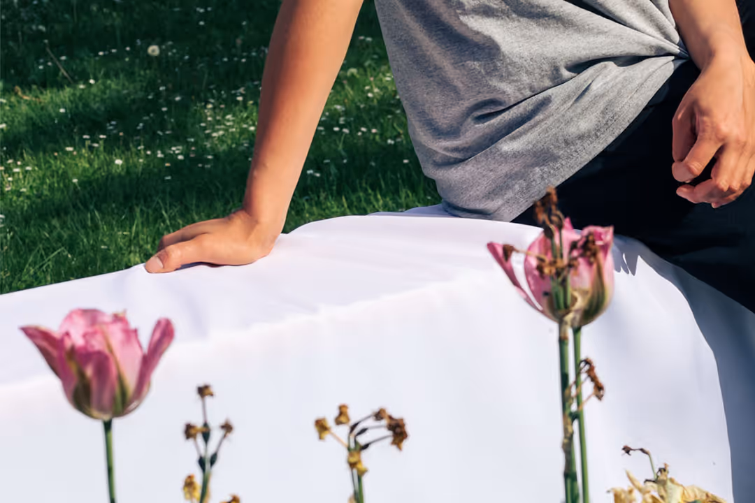 Bild einer Person, die auf einer Bierbank mit weißer, übergezogener Bierbankhusse sitzt. Die Bierbank steht auf einer grünen Wiese, im Vordergrund sind Blumen.