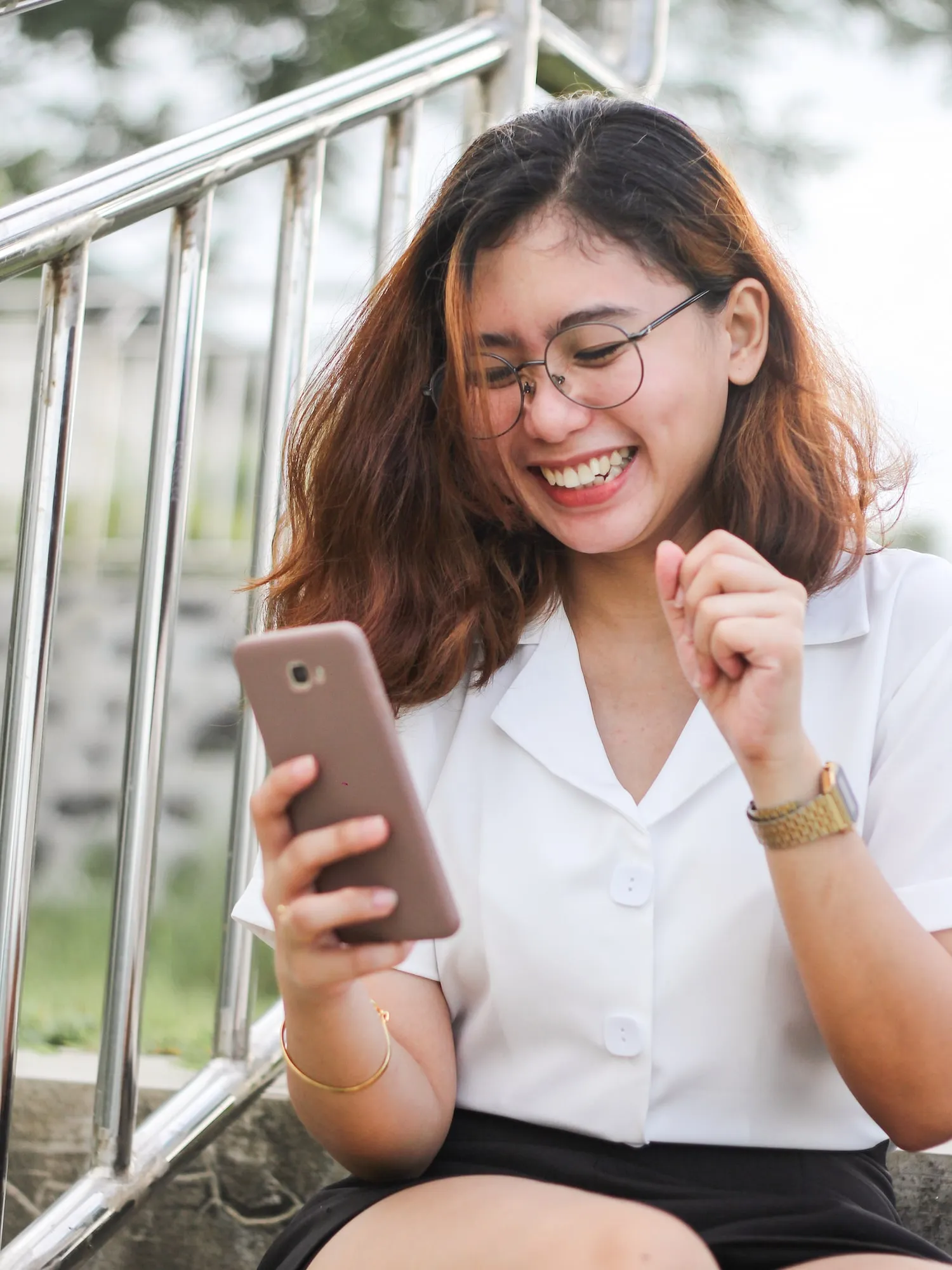 Smiling woman with glasses holding a smartphone and sitting near metal railings outdoors.