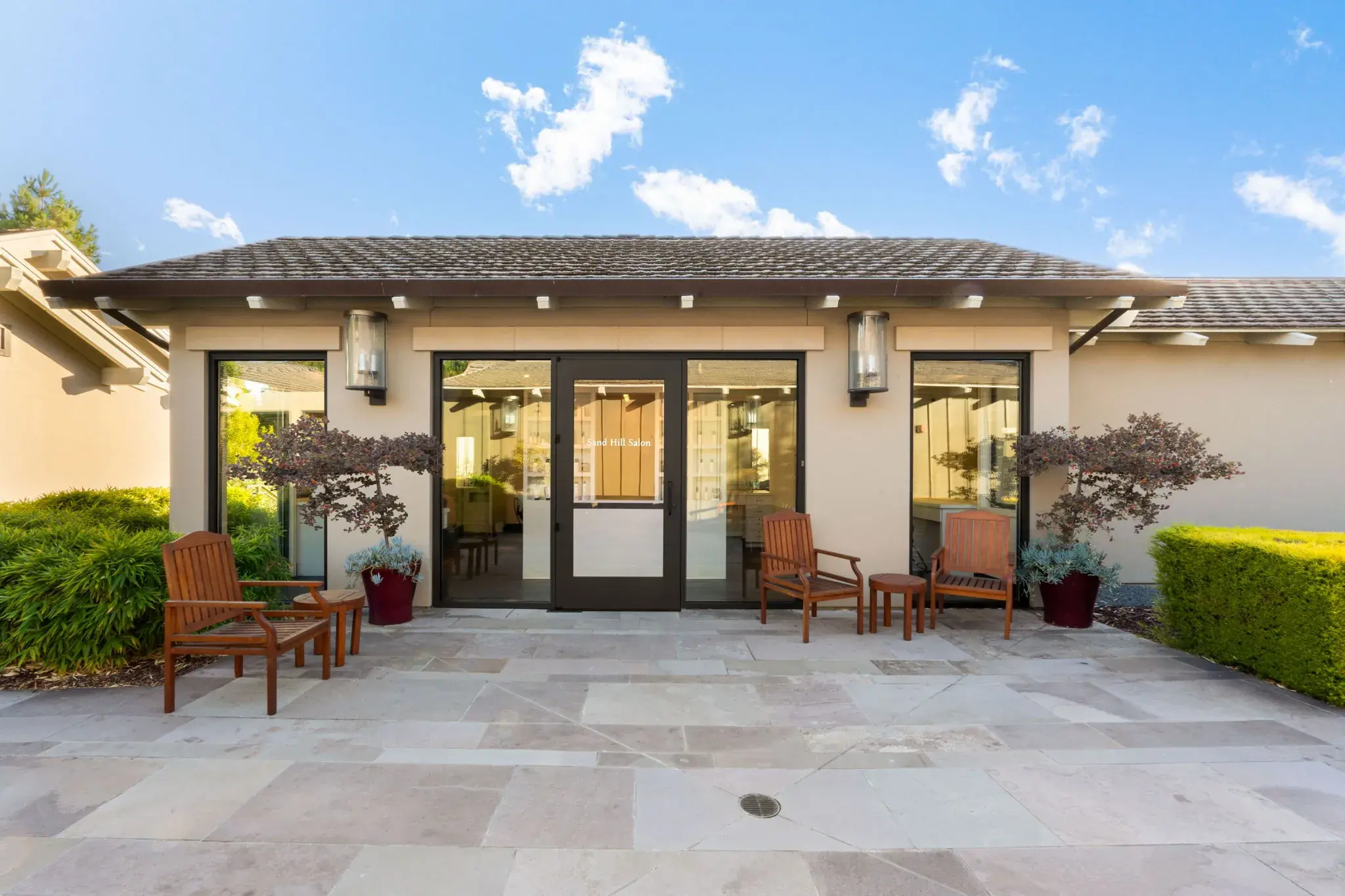 Patio outside a building with glass double doors, four wooden chairs, two side tables, and potted plants.