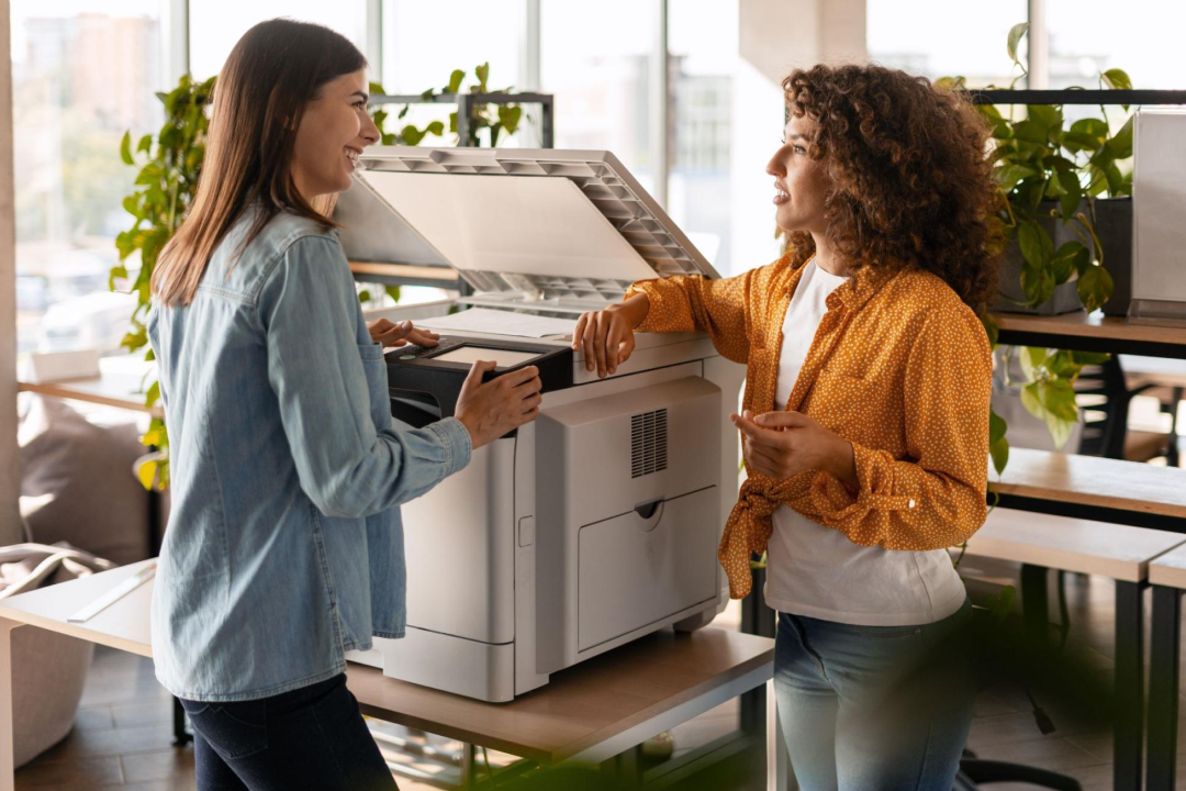 Two women in an office chat at the workplace printer.