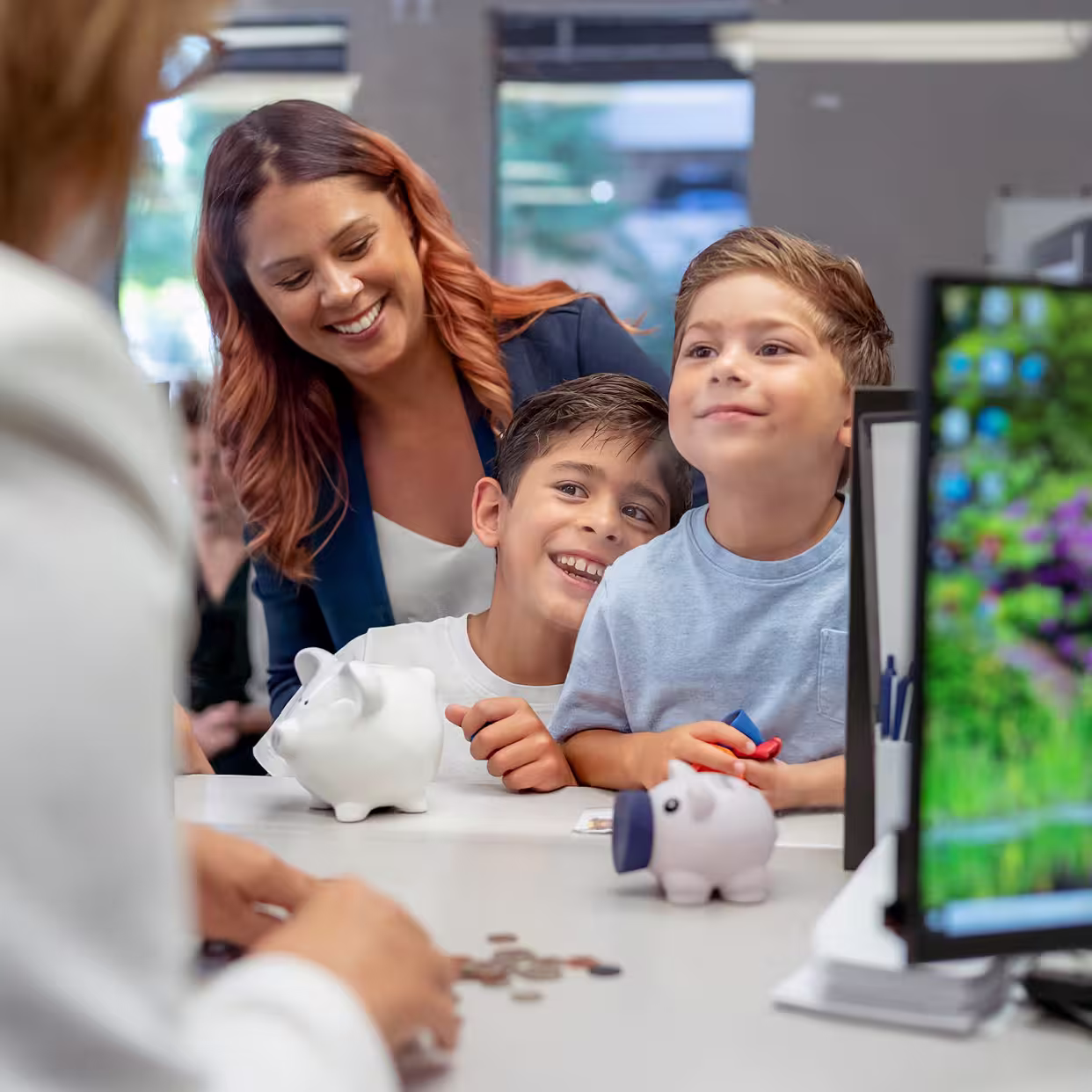A smiling woman with two young boys at a counter, with white and pig-shaped piggy banks and coins on the surface, in a bright indoor setting.