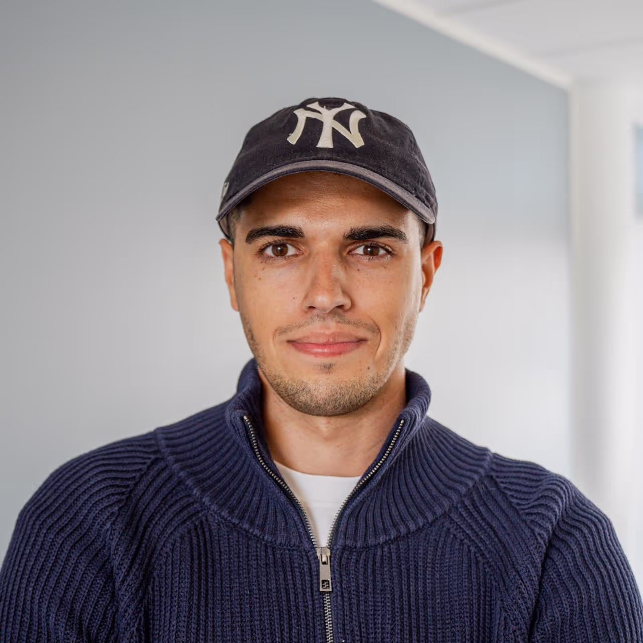 Young man wearing a navy blue zip-up sweater and a black baseball cap with a white logo, smiling slightly.