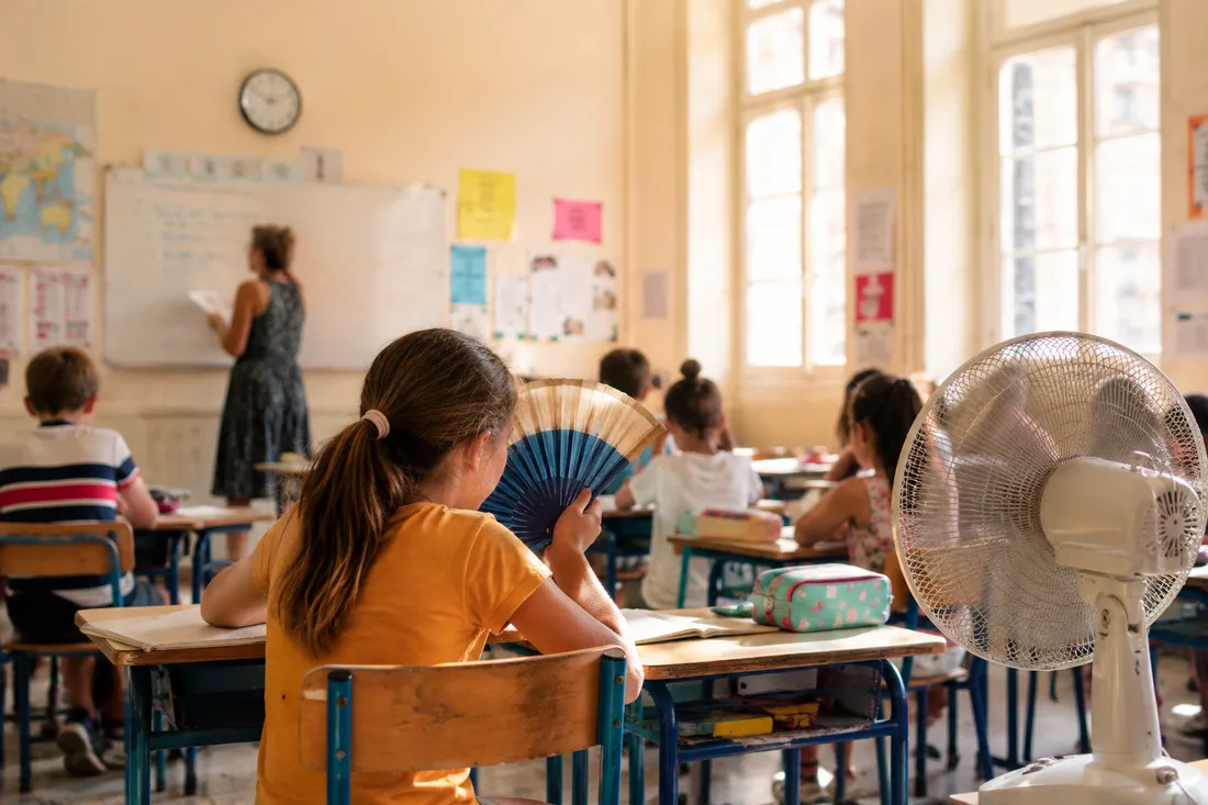 Élève s'éventant dans une salle de classe surchauffée, ventilateur sur pied au premier plan