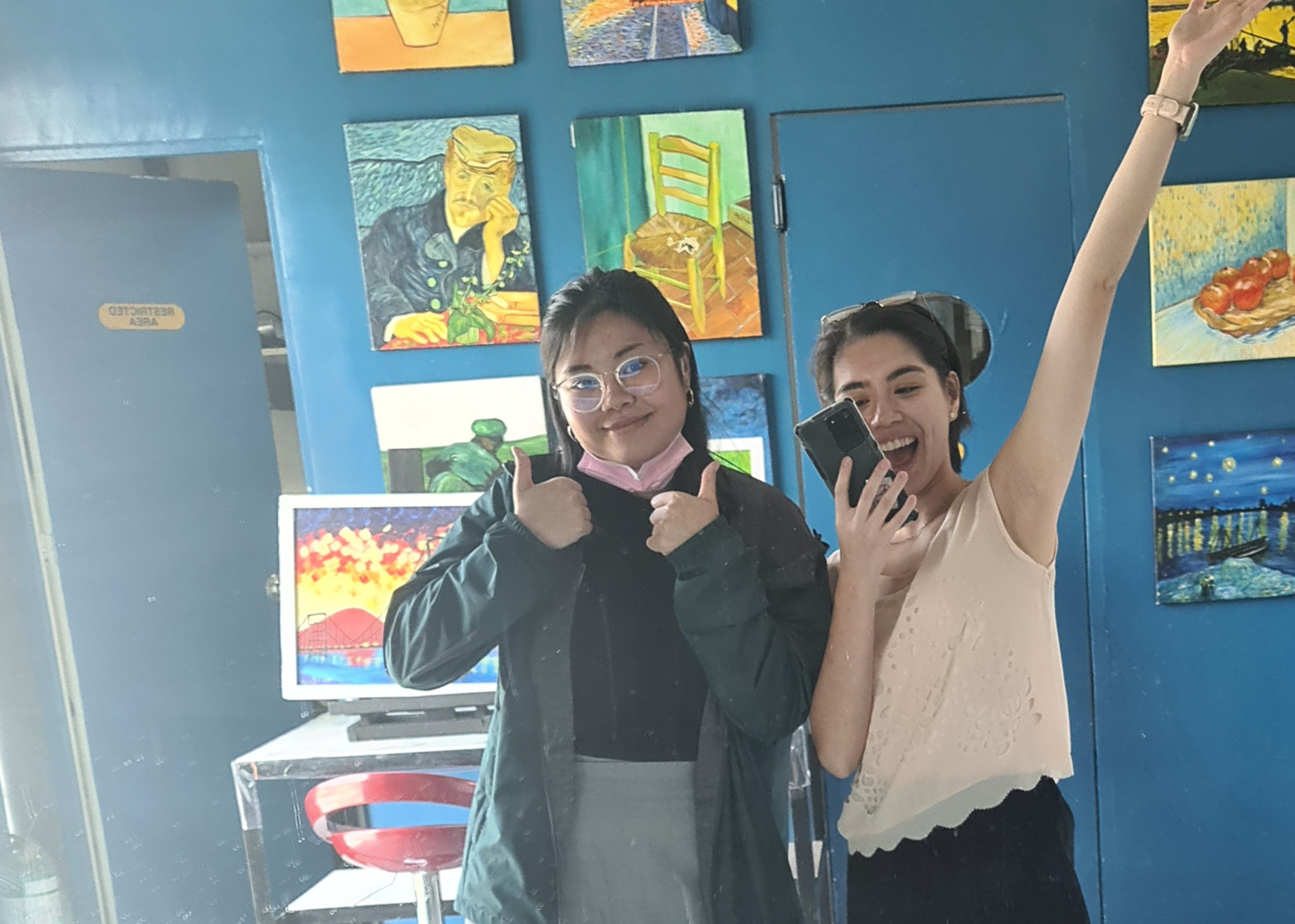 Dani and Liz in front of a blue wall looking very excited at their Sip N' Paint outing.