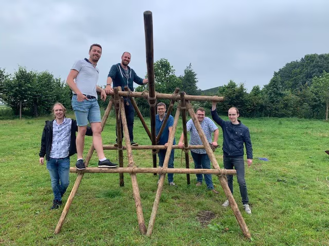 Twee mannen staan op de zelfgemaakte katapult van palen en touwen. Vier mannen staan op de grond ernaast
