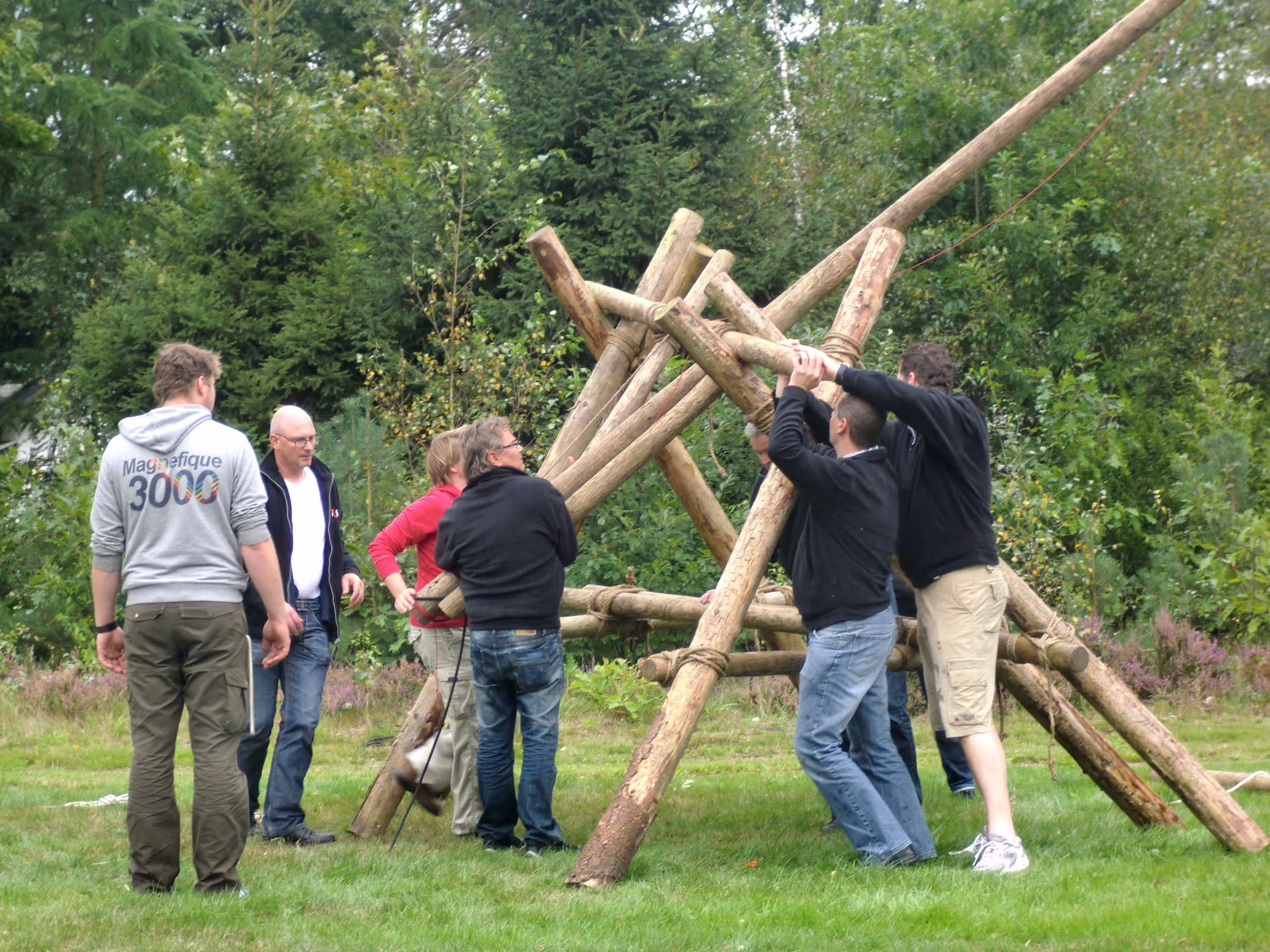 Circa zeven personen staan op een grasveld en schieten met een zelfgemaakte katapult van palen en touwen