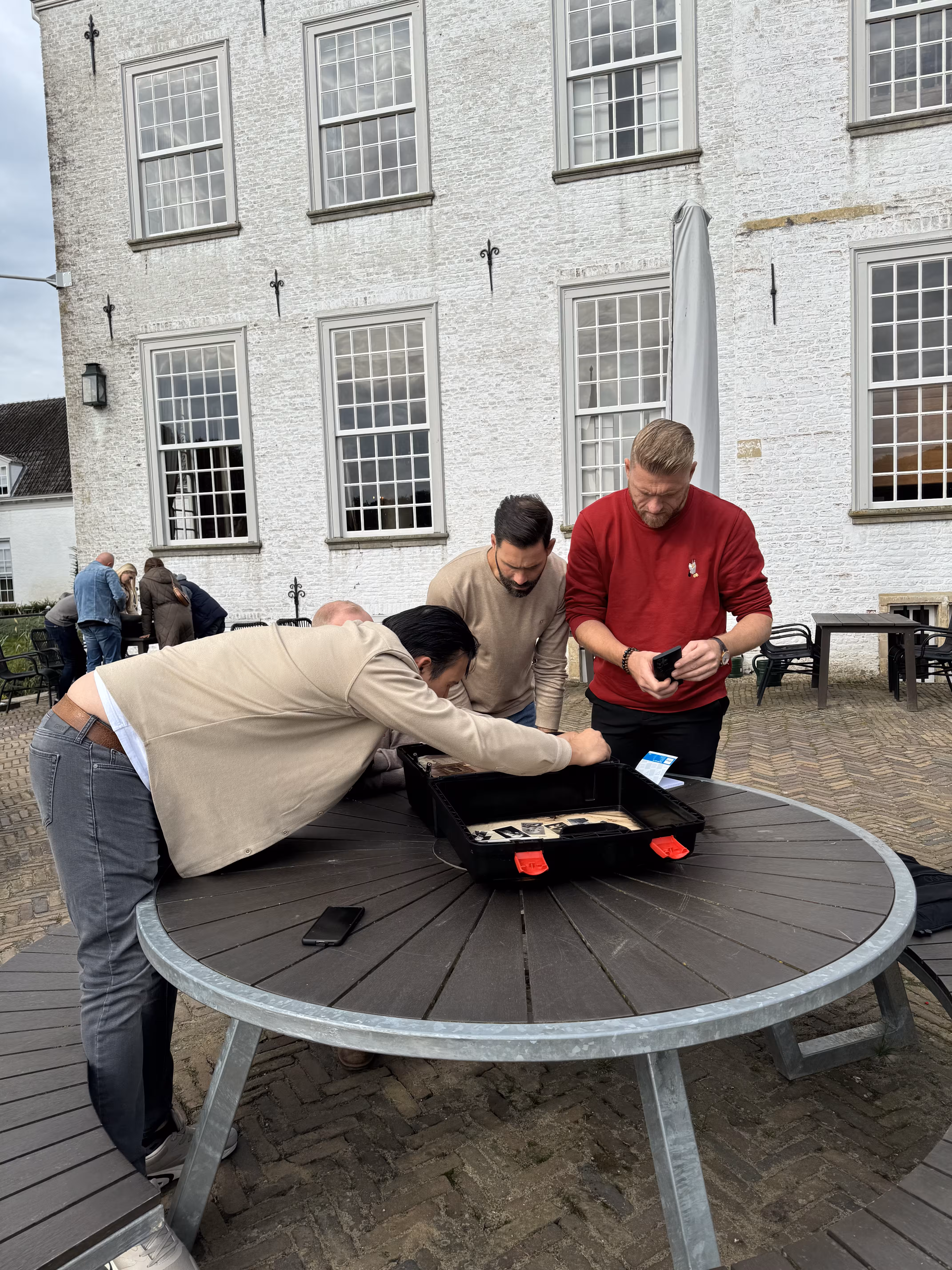 Drie mannen zitten buiten aan een ronde tafel bij een wit gebouw. Een man staat voorovergebogen 