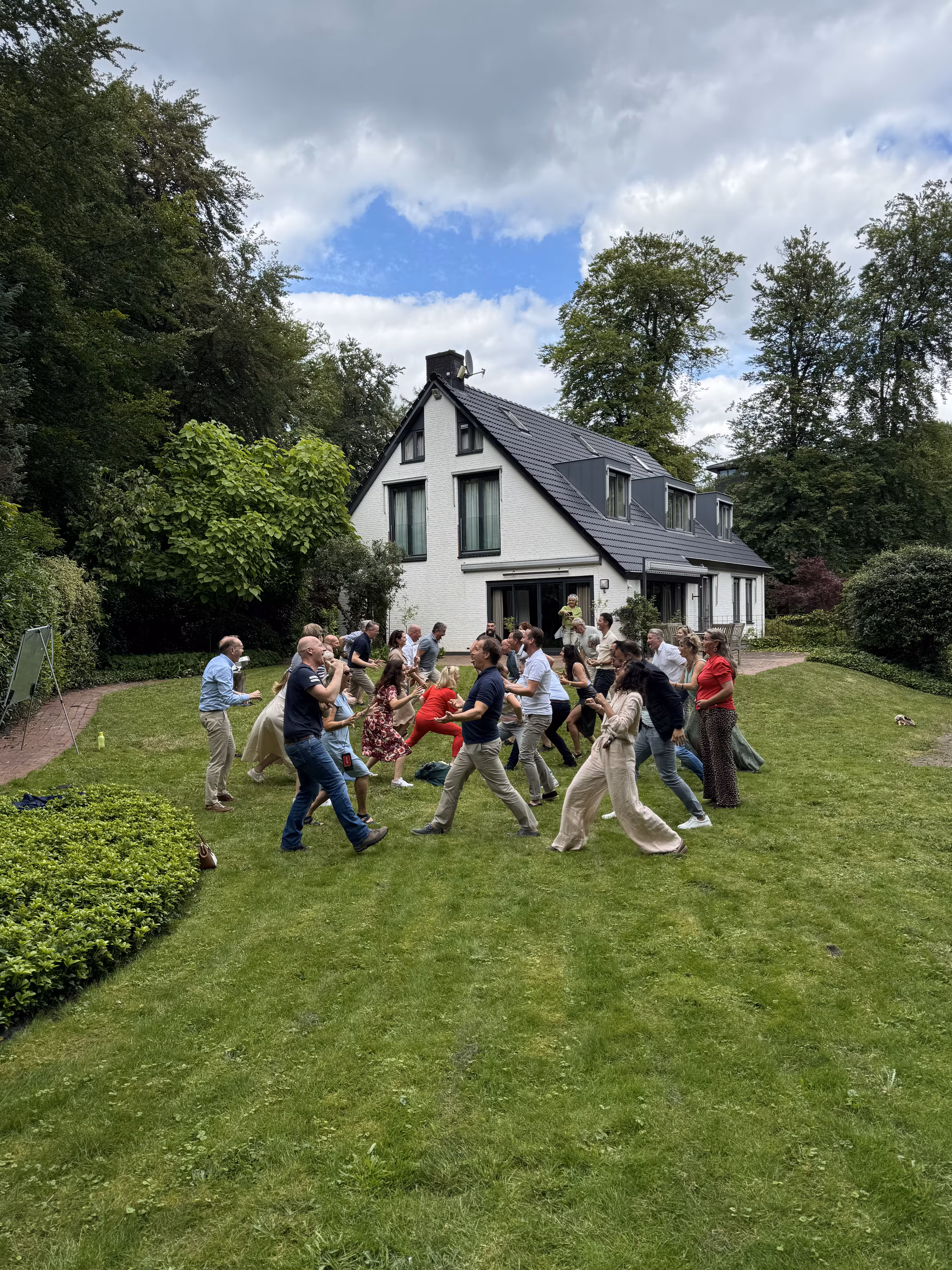 Op een grasveld bij een wit gebouw staan mensen tegenover elkaar en zetten een pas voorwaarts tijdens een HAKA workshop