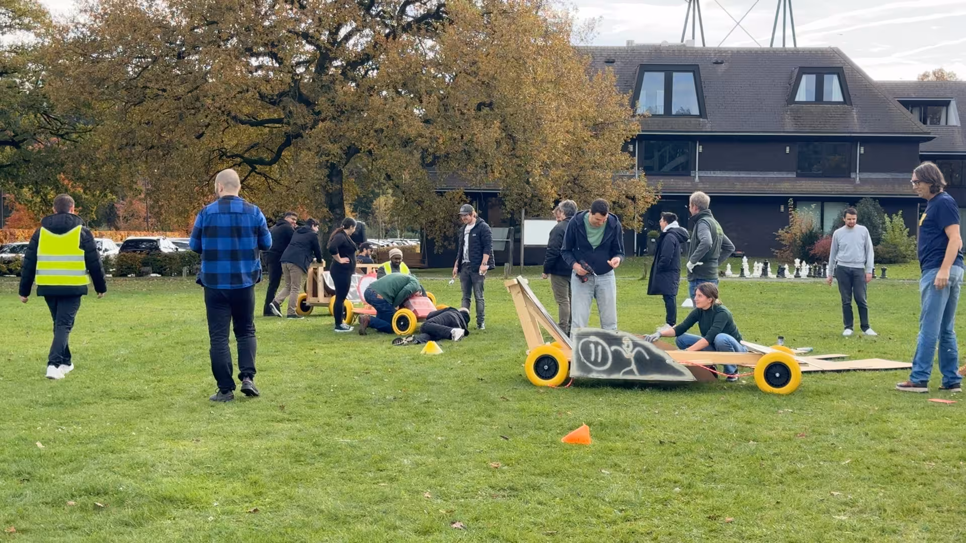 Drie teams werken aan een Formule 1 wagen op een grasveld met oranje en gele pionnen