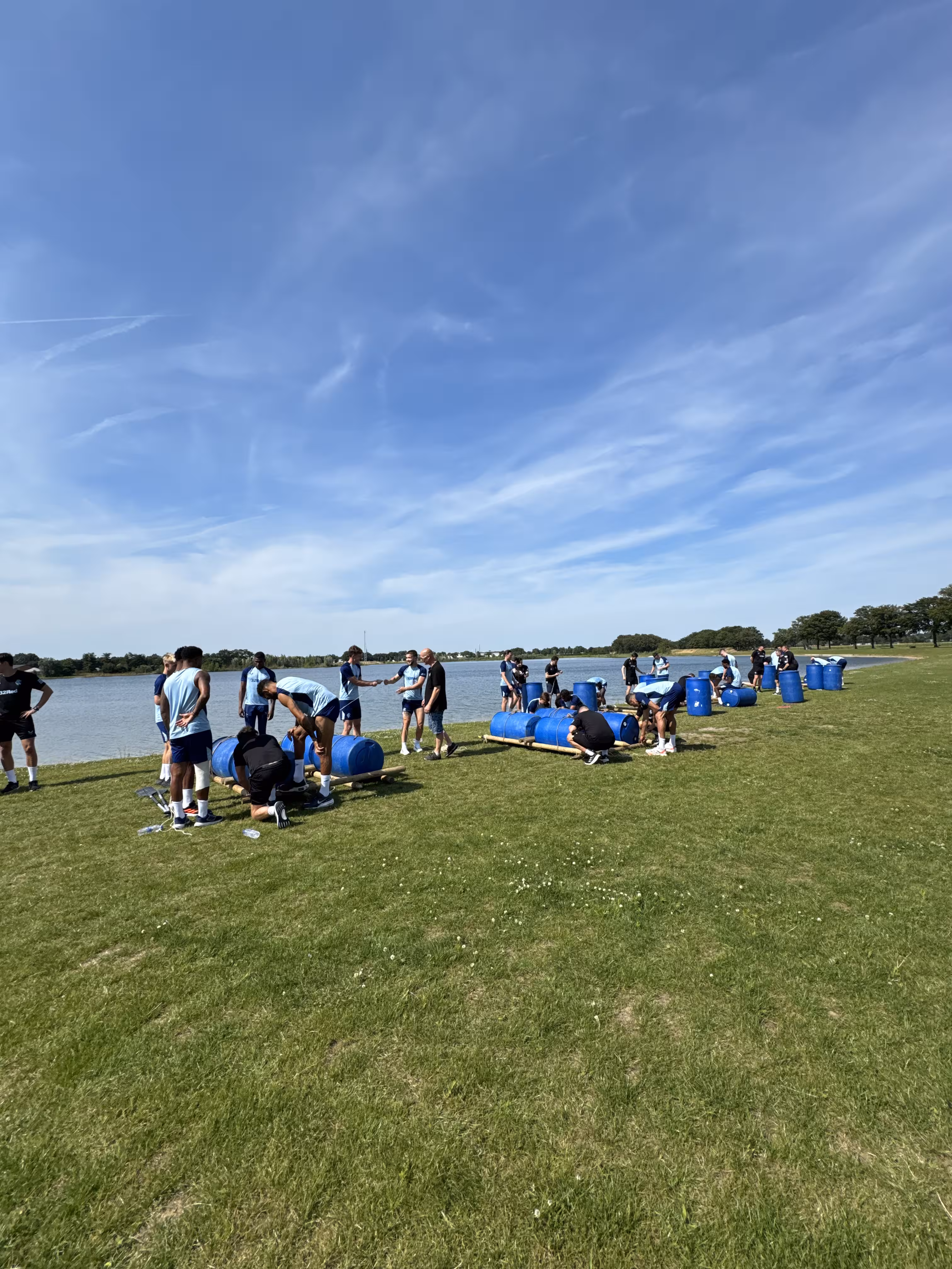 Op een grasveld bij een meer bouwen meerdere teams in lichtblauwe shirts aan een vlot