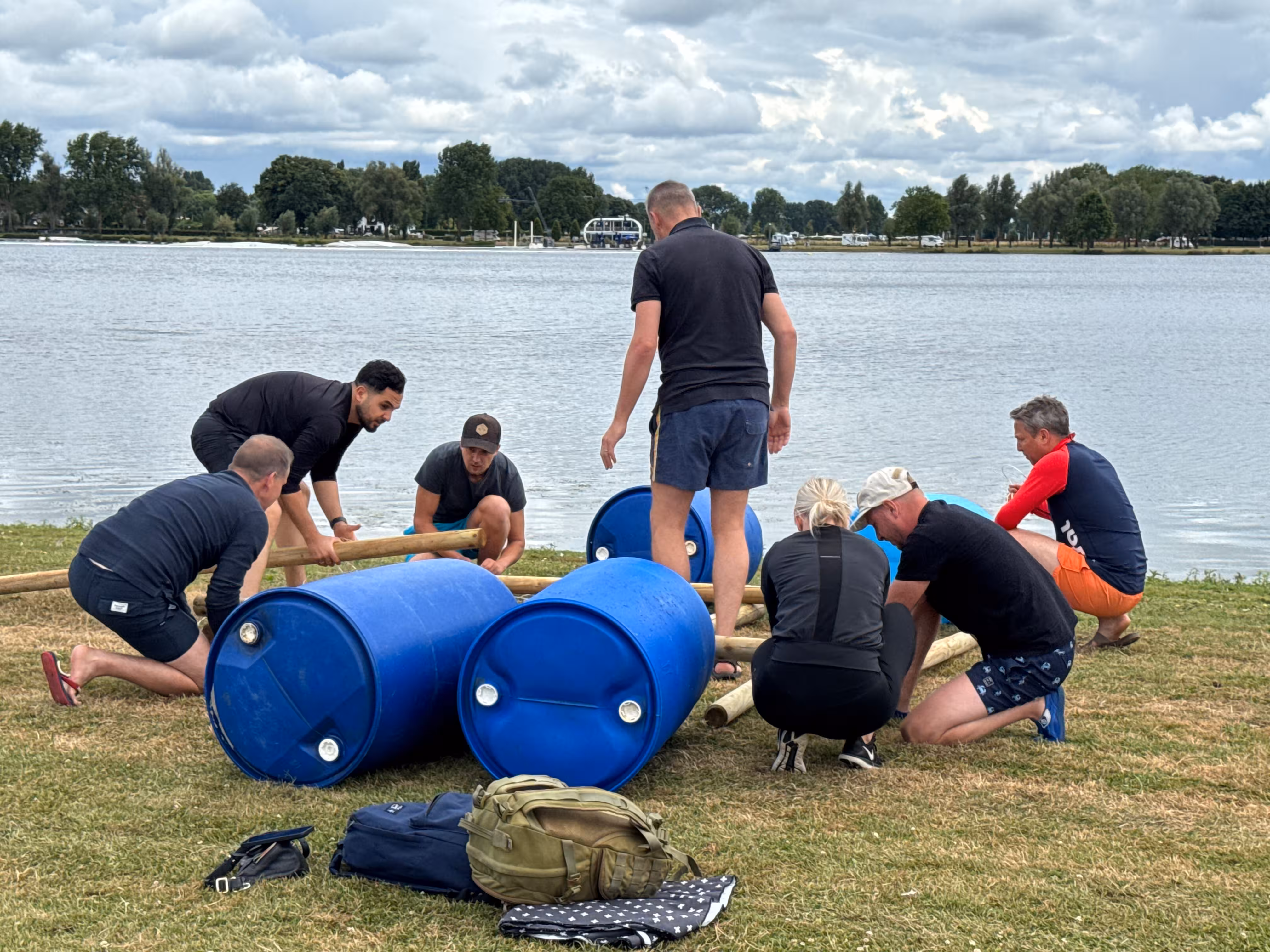 Zeven personen werken gezamenlijk aan het bouwen van een constructie met houten palen en touwen. Ze nemen deel aan de workshop Vlot Bouwen
