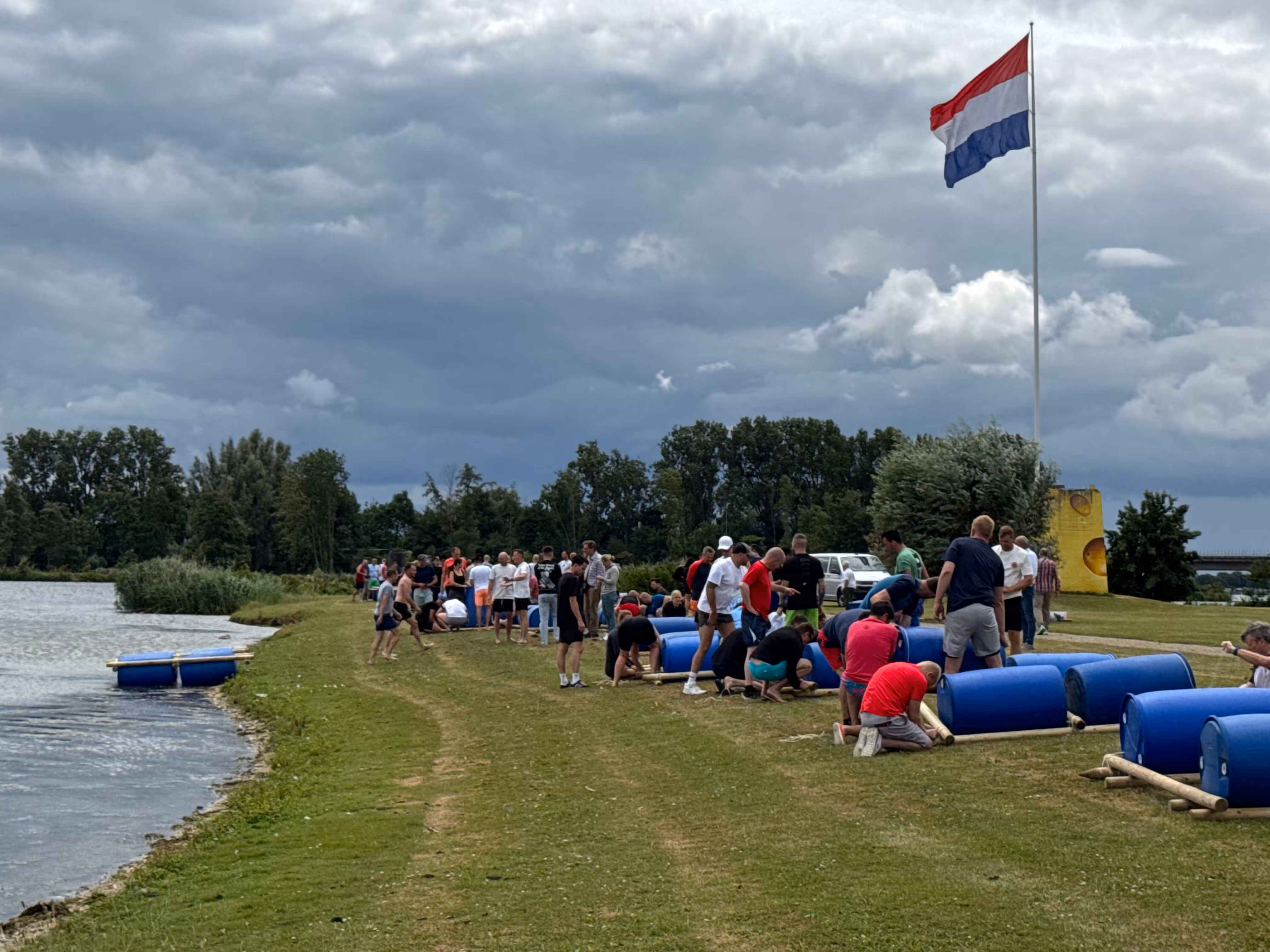 Op een bewolkte dag wordt er op een grasveld met een Nederlandse vlag door teams gewerkt aan het bouwen van een vlot.